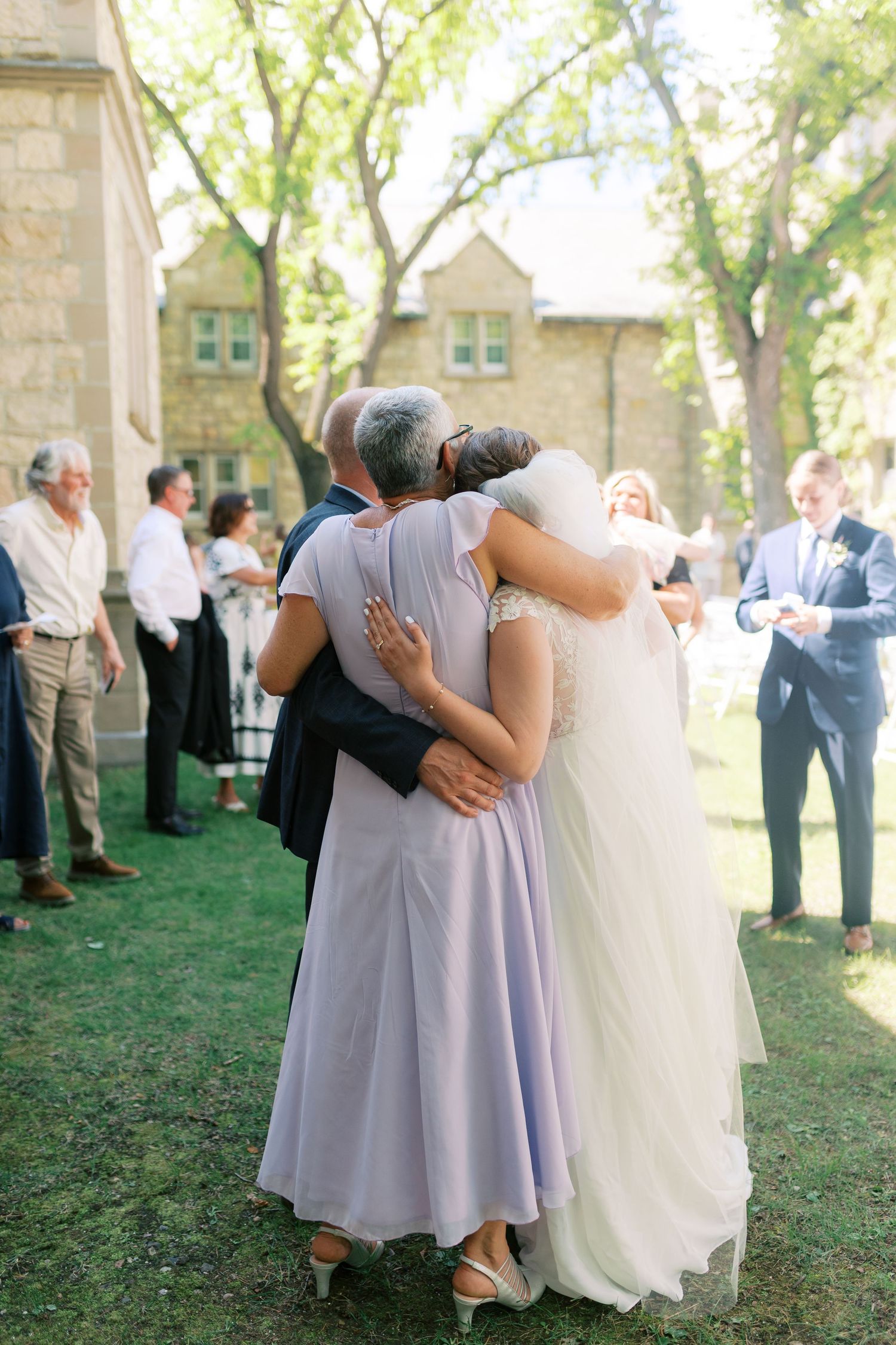 Bride hugging parents from behind at outdoor wedding reception on green lawn near stone building.