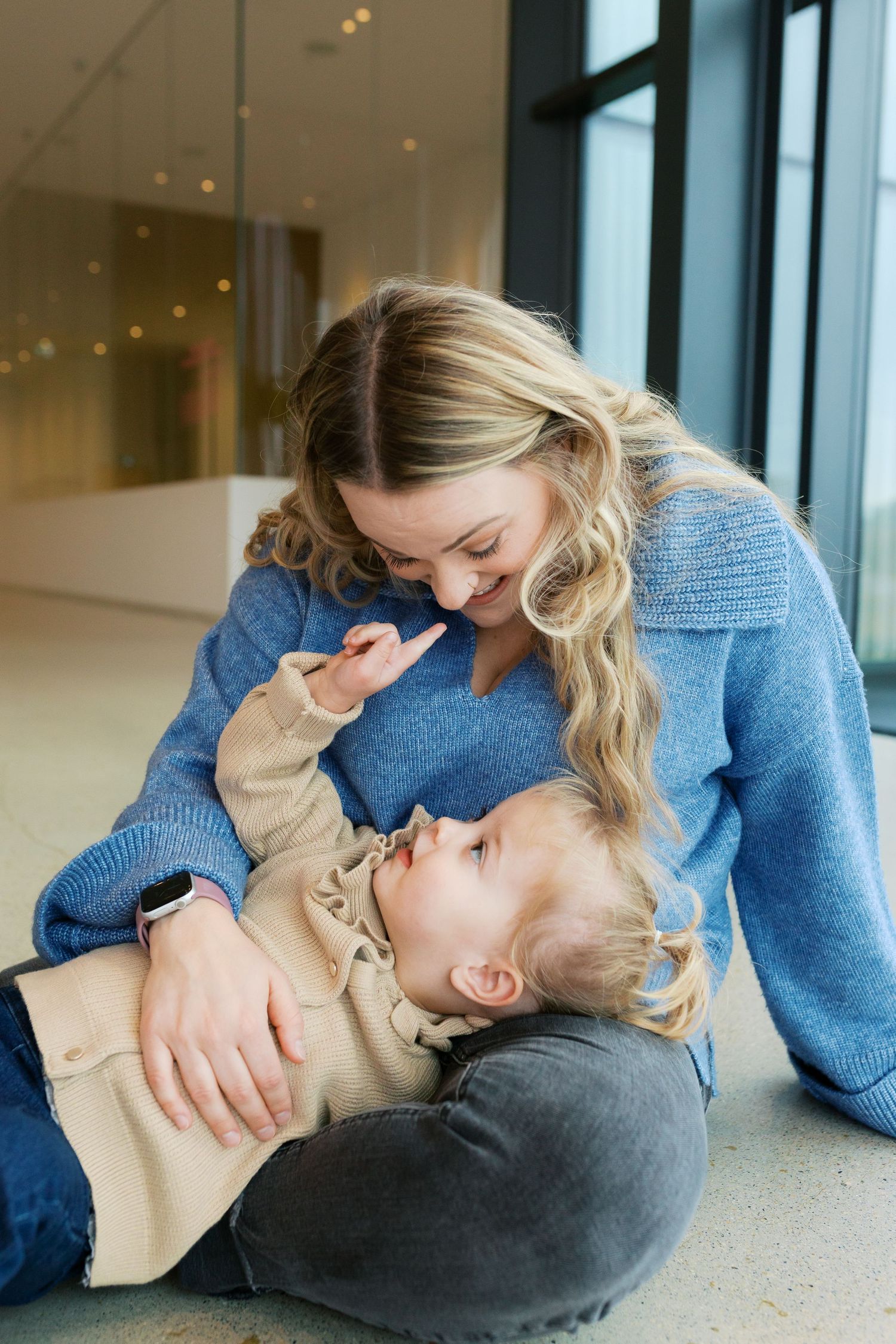 Parent and child share playful moment while lying on floor near large windows.