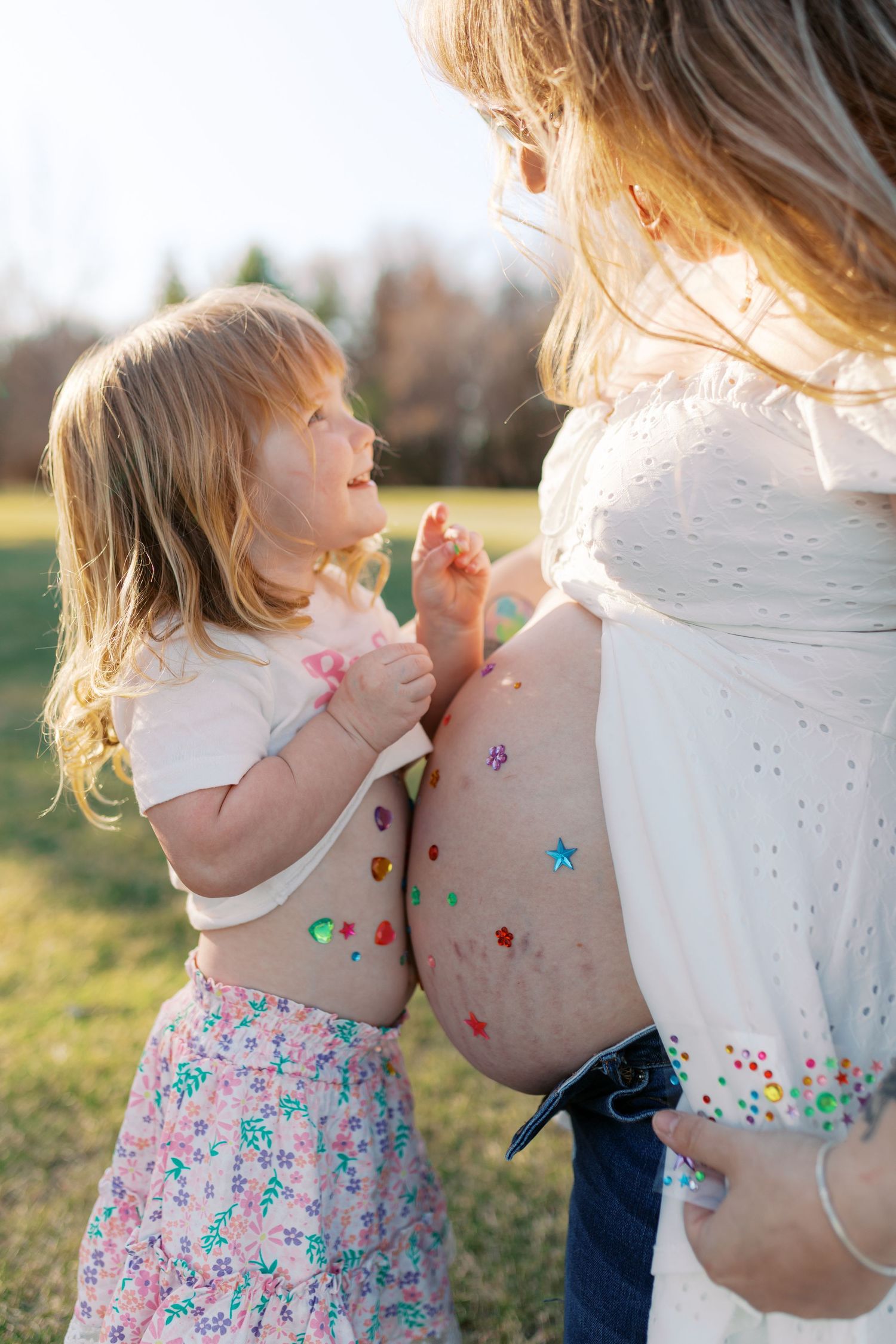 A young girl decorates her pregnant mother's baby bump with colorful stickers outdoors in a sunny park.
