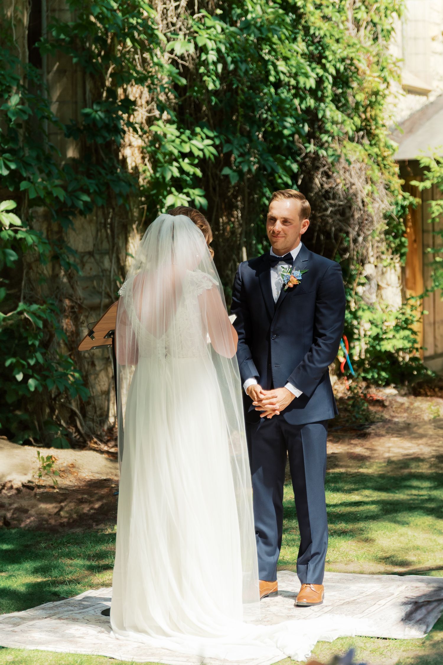 Groom in navy suit smiles at bride in white wedding gown and veil during outdoor ceremony.