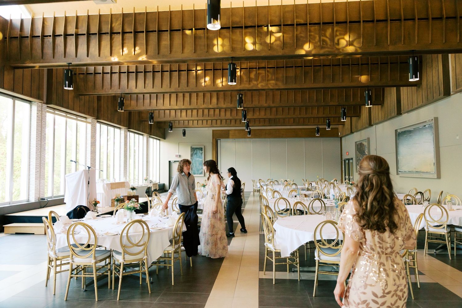Wedding reception venue setup with round tables, gold chairs, and large windows in a modern wood-paneled banquet hall.
