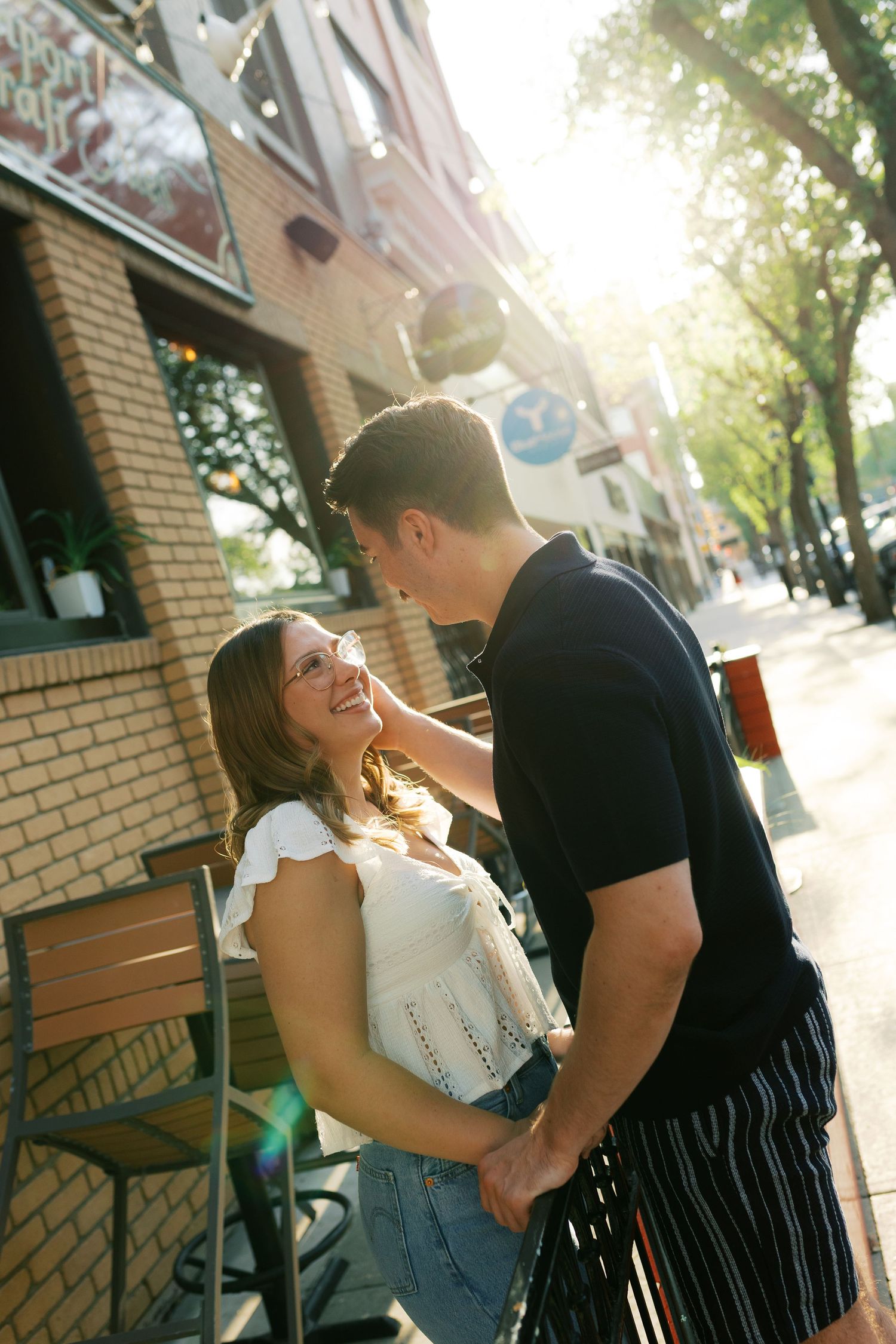 A young couple holds hands and smiles at each other on a sunny urban sidewalk outside a brick building.