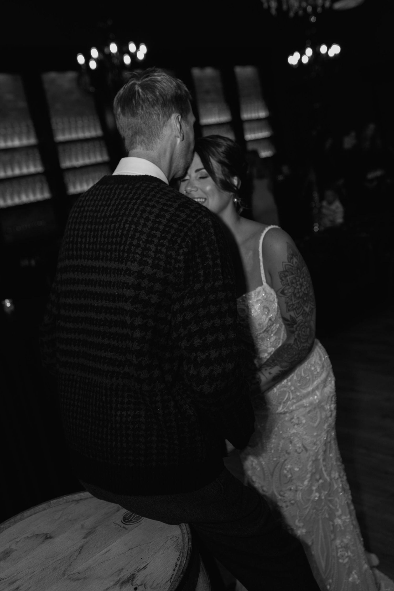 Black and white photo of a bride in lace gown leaning on groom in checkered jacket during romantic wedding dance.