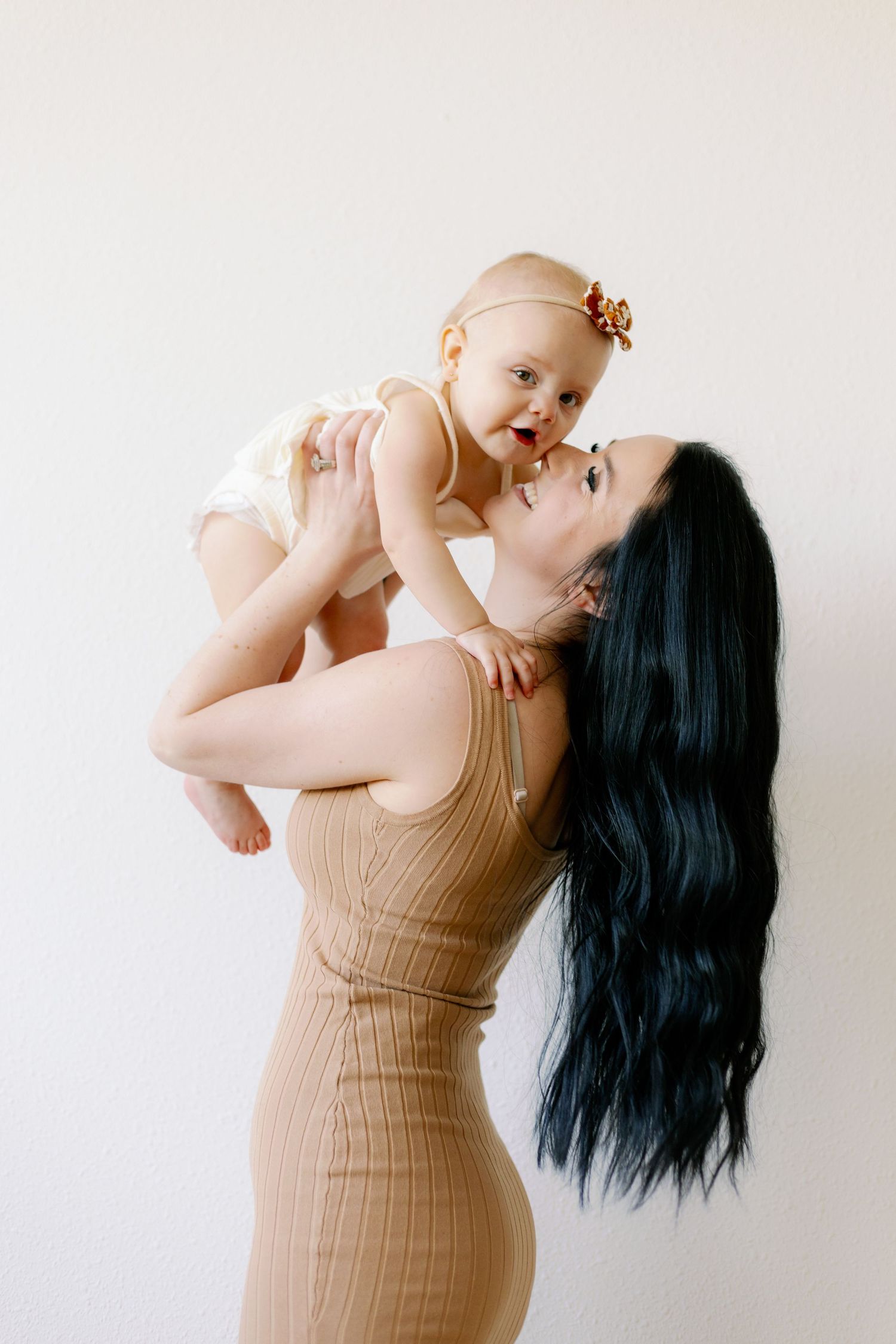 Parent lifts smiling baby in white onesie against neutral background.