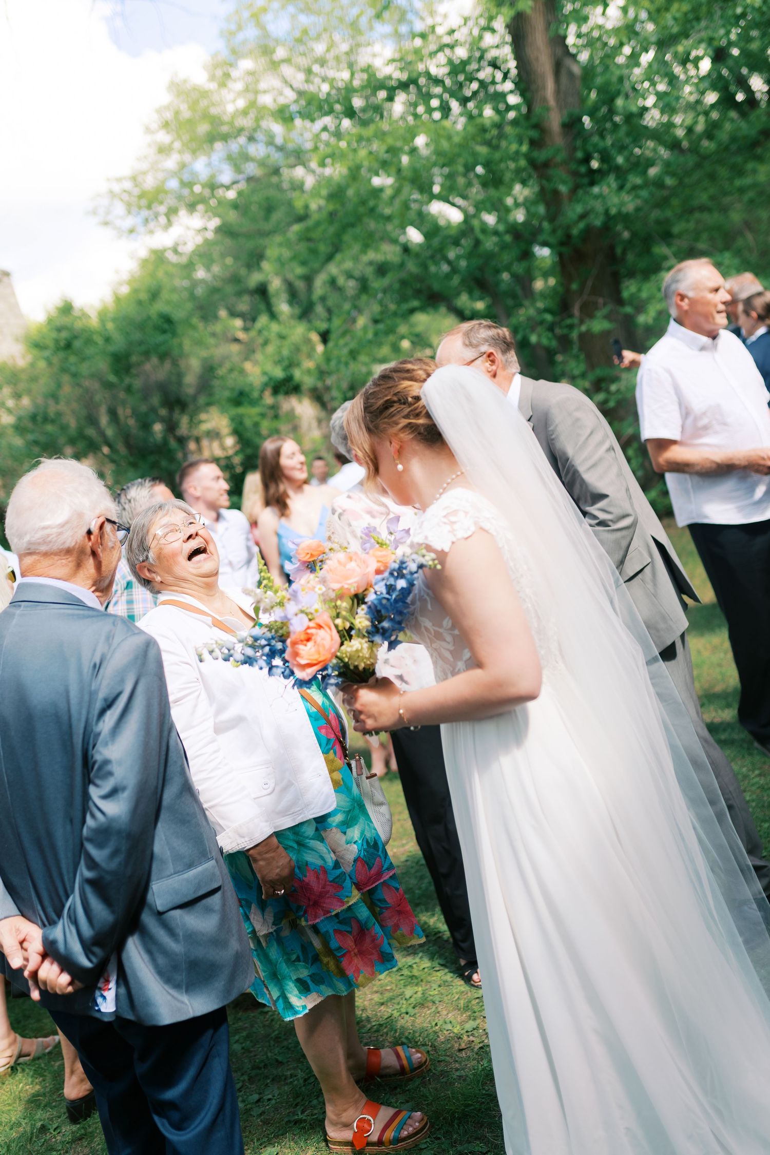 A smiling bride holding a colorful bouquet greets laughing guests outdoors at a summer wedding reception.