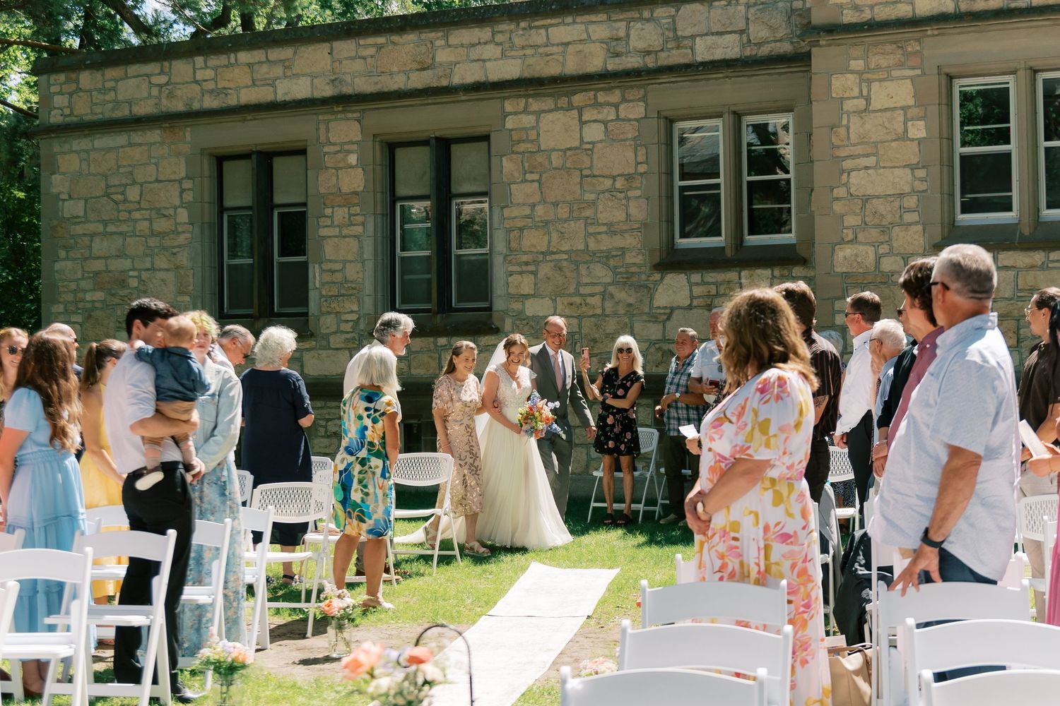 Outdoor wedding ceremony on sunny day with guests seated in white chairs on green lawn beside stone building.