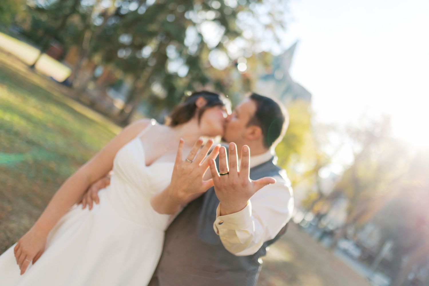 Newlywed couple kissing and showing off wedding rings outdoors in soft sunlight on their wedding day.