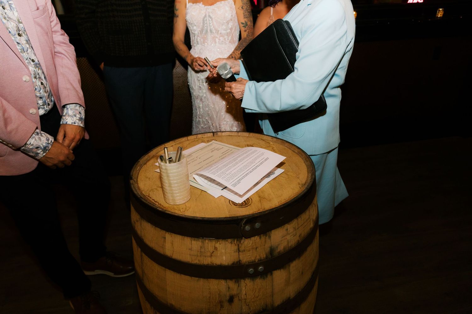 Outdoor wedding ceremony at a rustic wine barrel altar with bride, groom, and officiant signing documents at night.