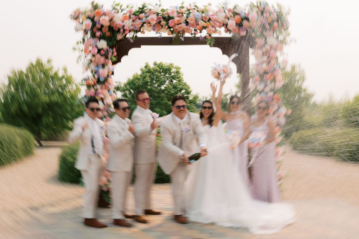 Wedding party celebrating under floral arch with pink blooms, bride and groom surrounded by groomsmen and bridesmaids outdoors.