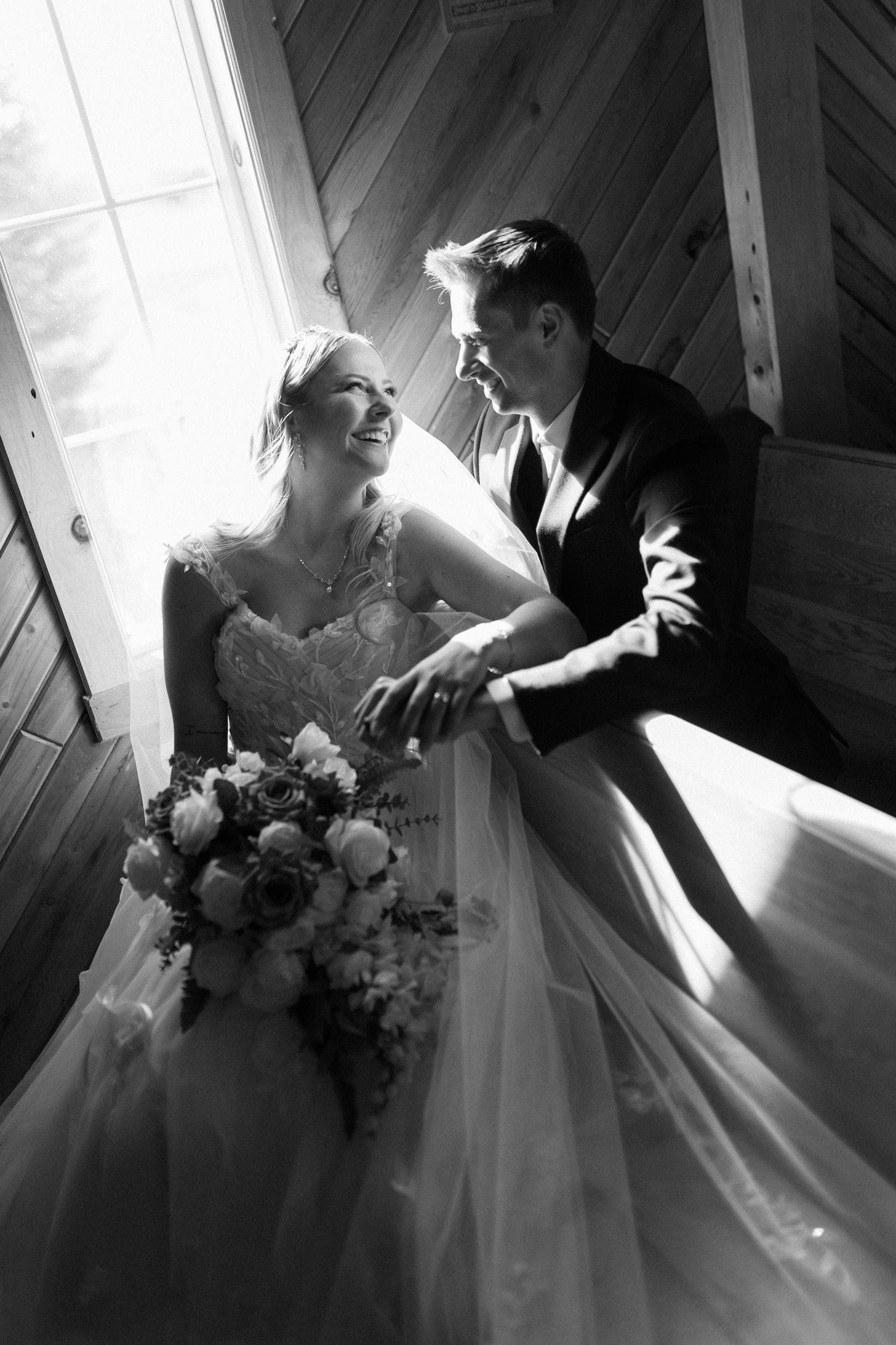 Black and white wedding photo of smiling bride holding bouquet and groom leaning on railing by window in wooden venue.