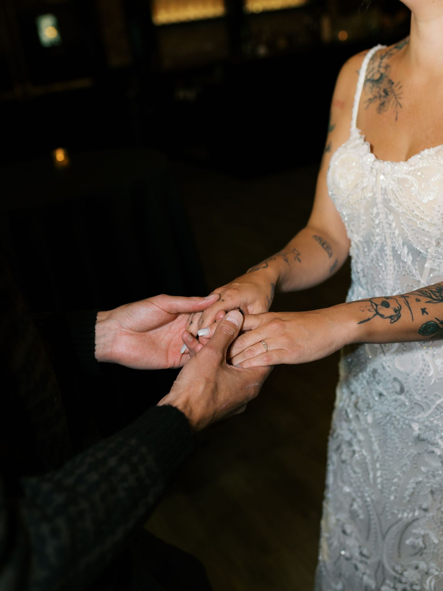 Bride in white lace gown with tattoos receiving wedding ring during ceremony.