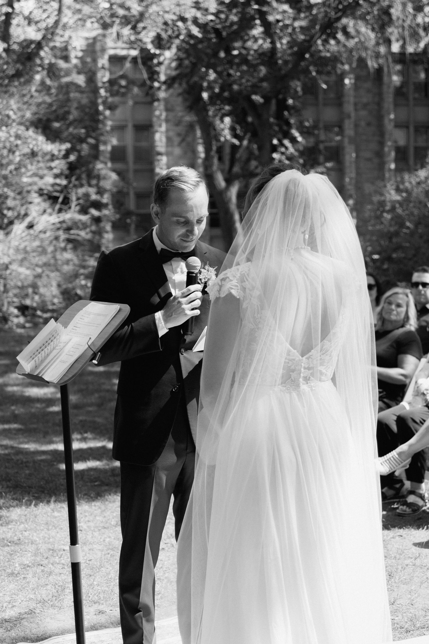 Black and white outdoor wedding ceremony with groom in tuxedo reading vows to bride in lace gown and veil.