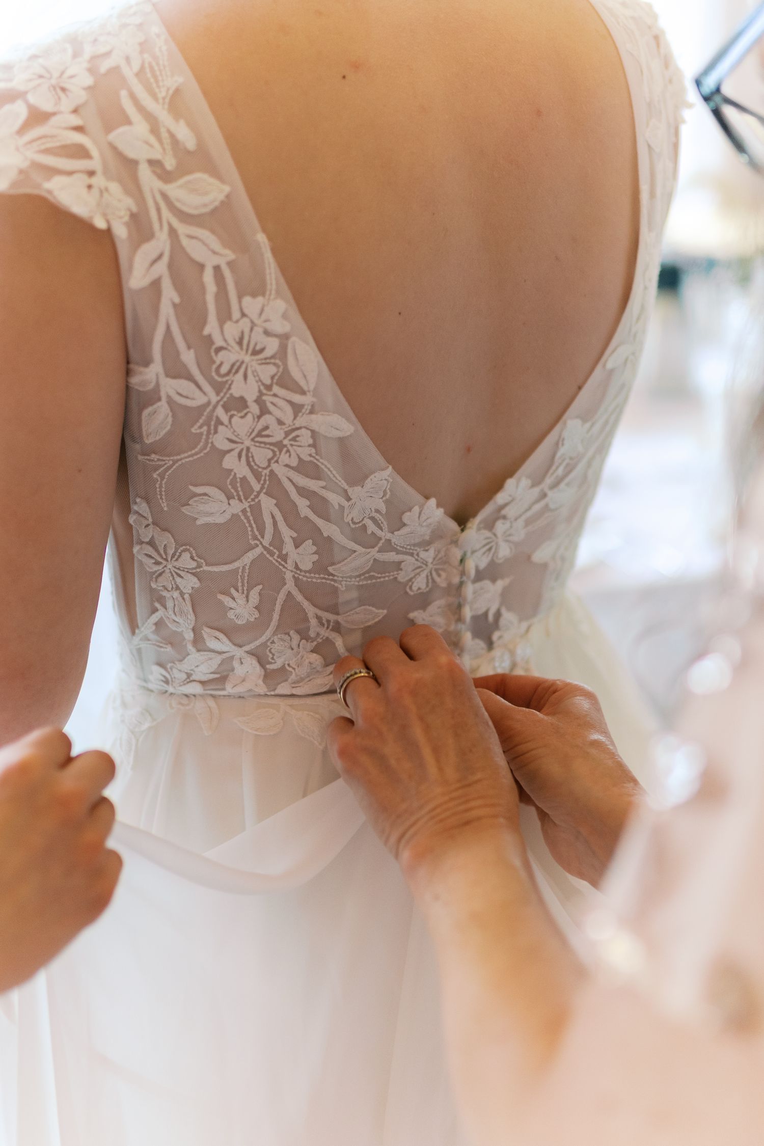 Close-up of intricate lace back wedding dress being buttoned by helping hands.
