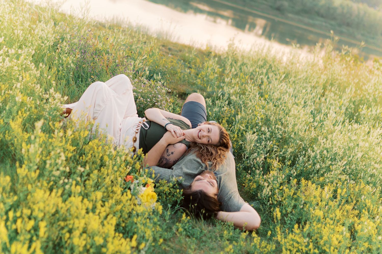 Couple lying in a sunny wildflower meadow by a lake, sharing a romantic moment among yellow blooms.