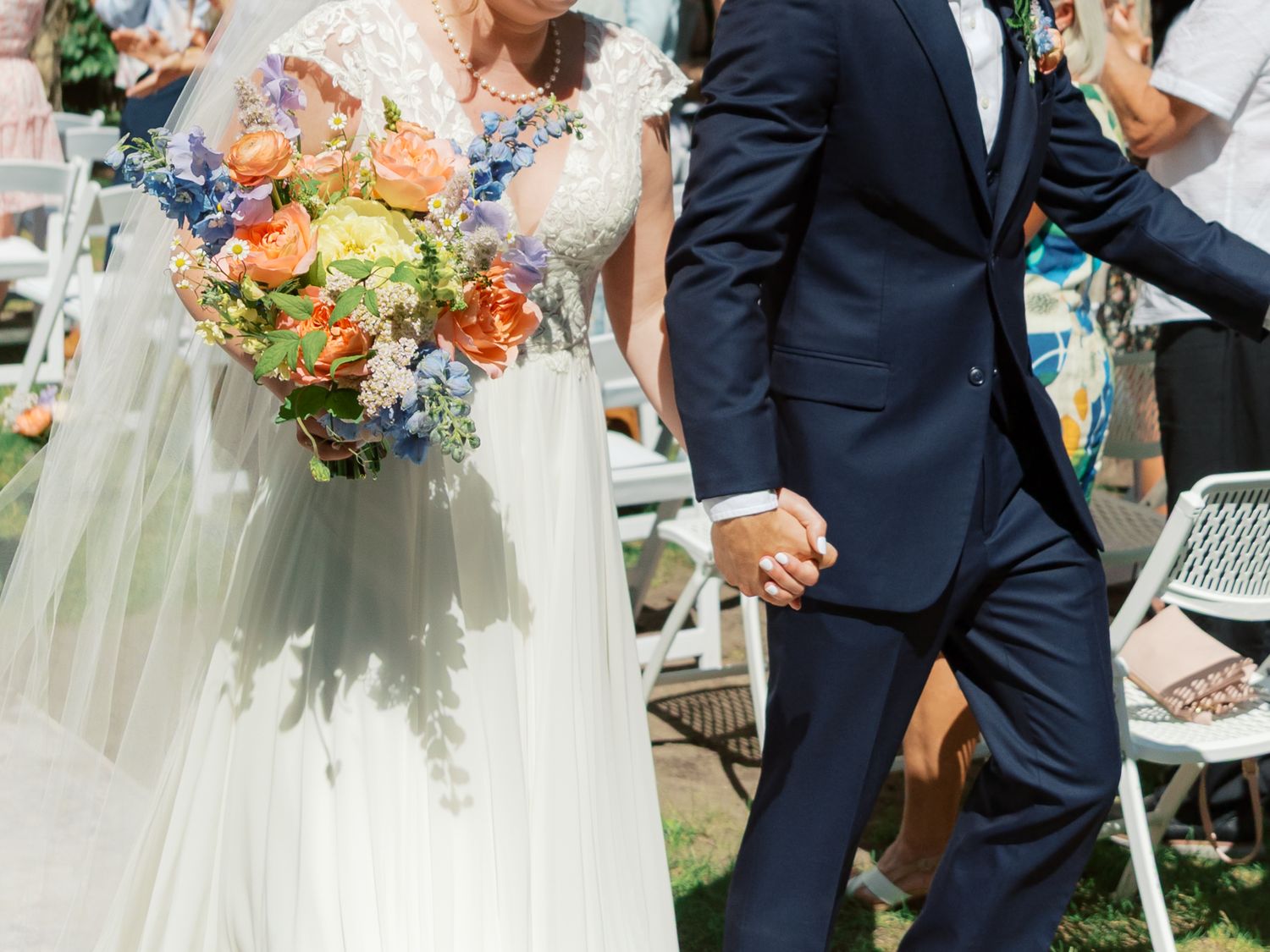 Bride in white gown holding colorful bouquet walks hand-in-hand with groom in navy suit at outdoor wedding ceremony.