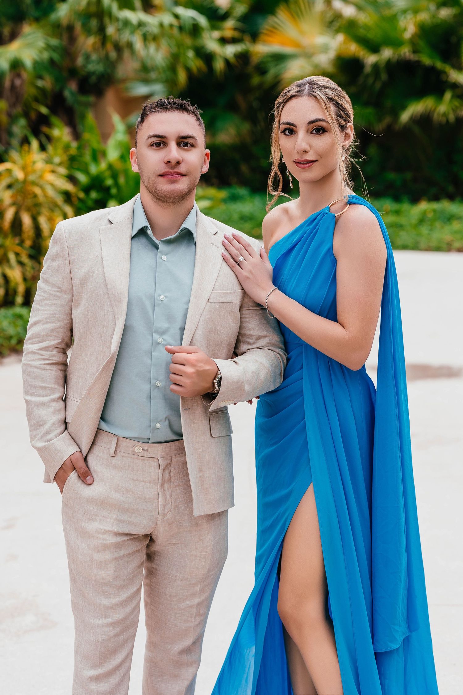 Formal couple poses against tropical foliage in beige suit and one-shoulder blue gown.