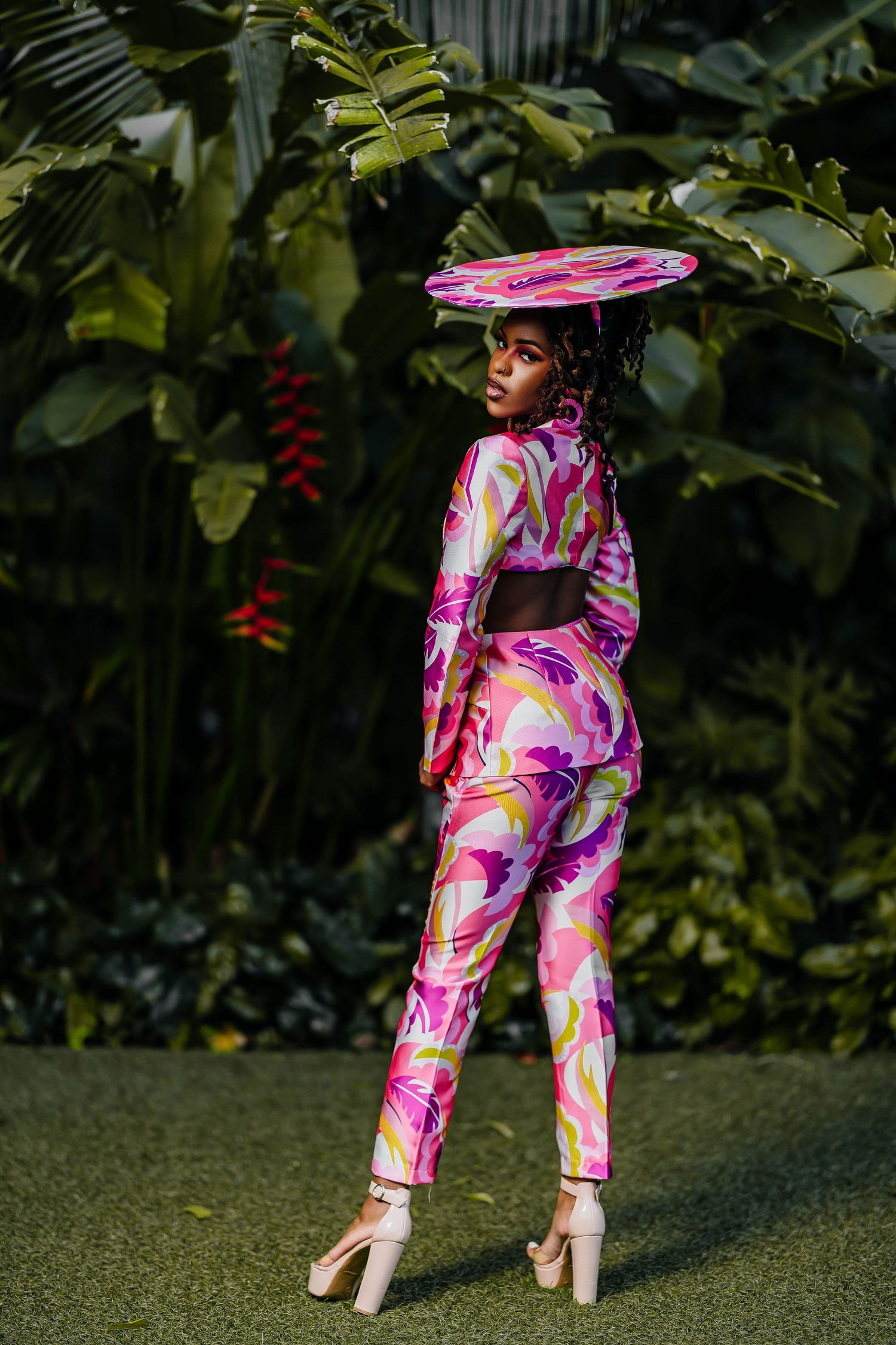 Modern fashion look featuring pink printed jumpsuit and matching disc hat against lush greenery.