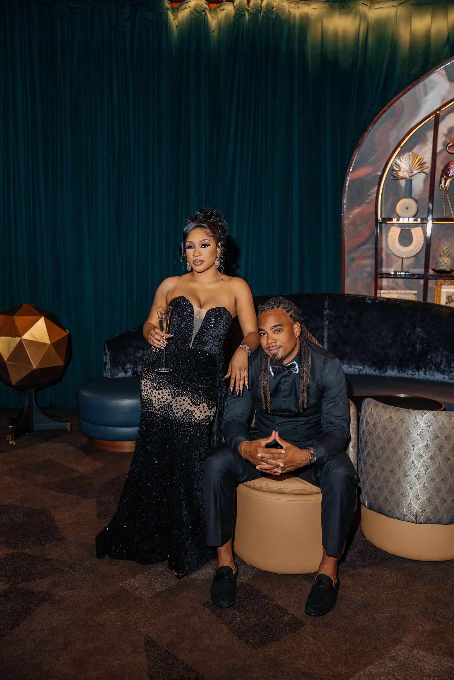 Couple in formal black attire poses for photos in an upscale venue with teal walls.