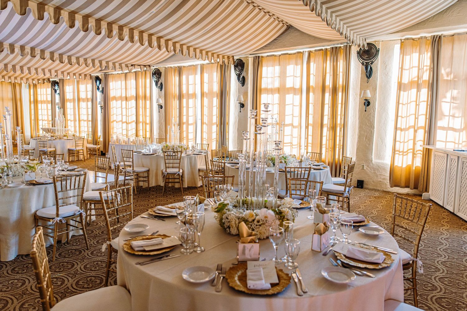 Elegant wedding reception hall with gold Chiavari chairs, white draped tables, floral centerpieces, and striped tent ceiling.