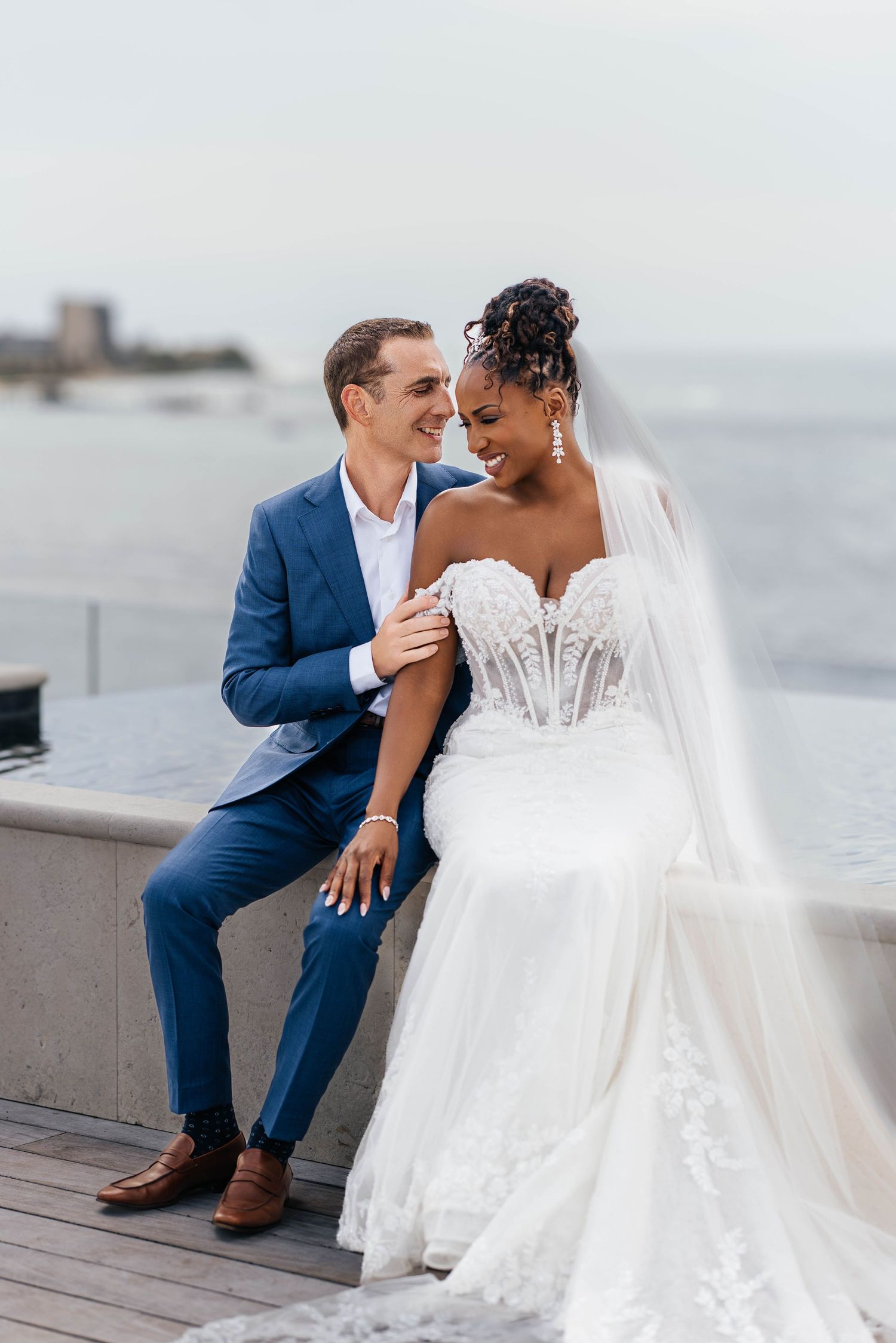 Wedding couple shares candid laughing moments while sitting together on seaside boardwalk.