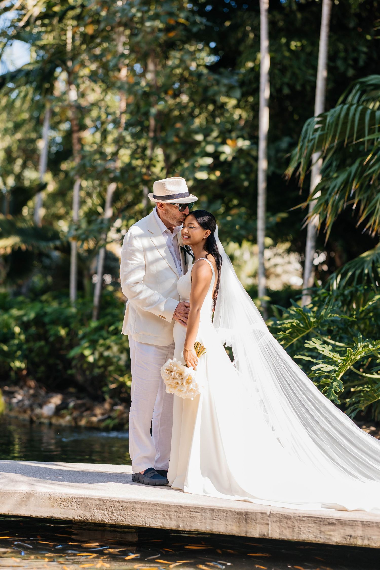 A romantic wedding couple shares tender kisses in a tropical garden setting with palm trees and natural lighting.