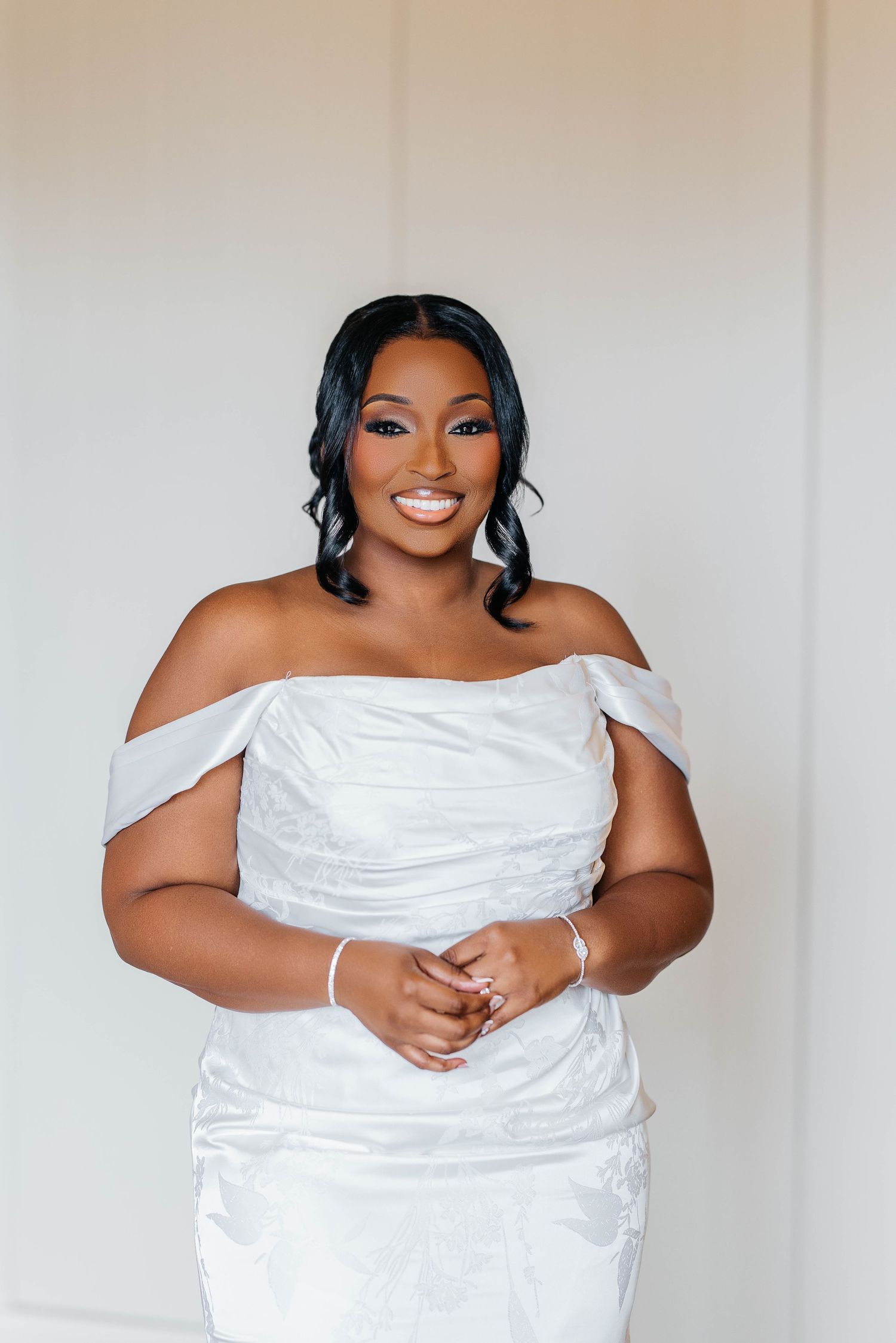 A smiling bride in an off-shoulder white gown with elegant makeup and dark wavy hair poses gracefully indoors.