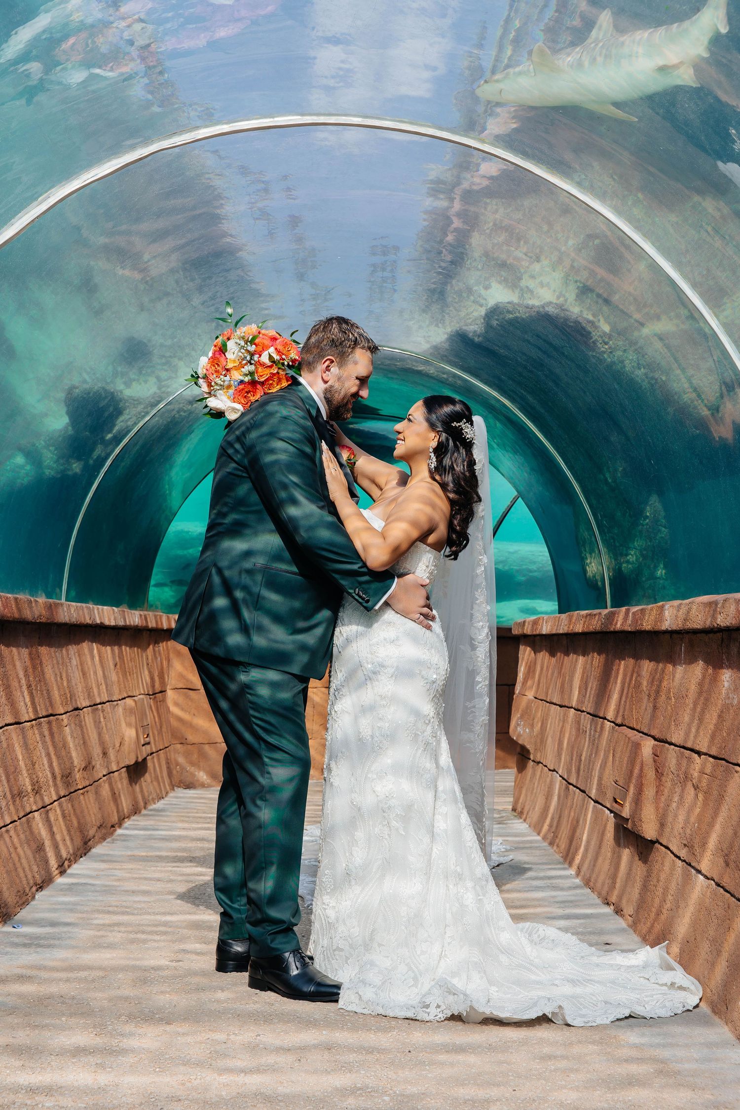 Dramatic wedding portrait in underwater viewing tunnel with flowing white dress and emerald suit.