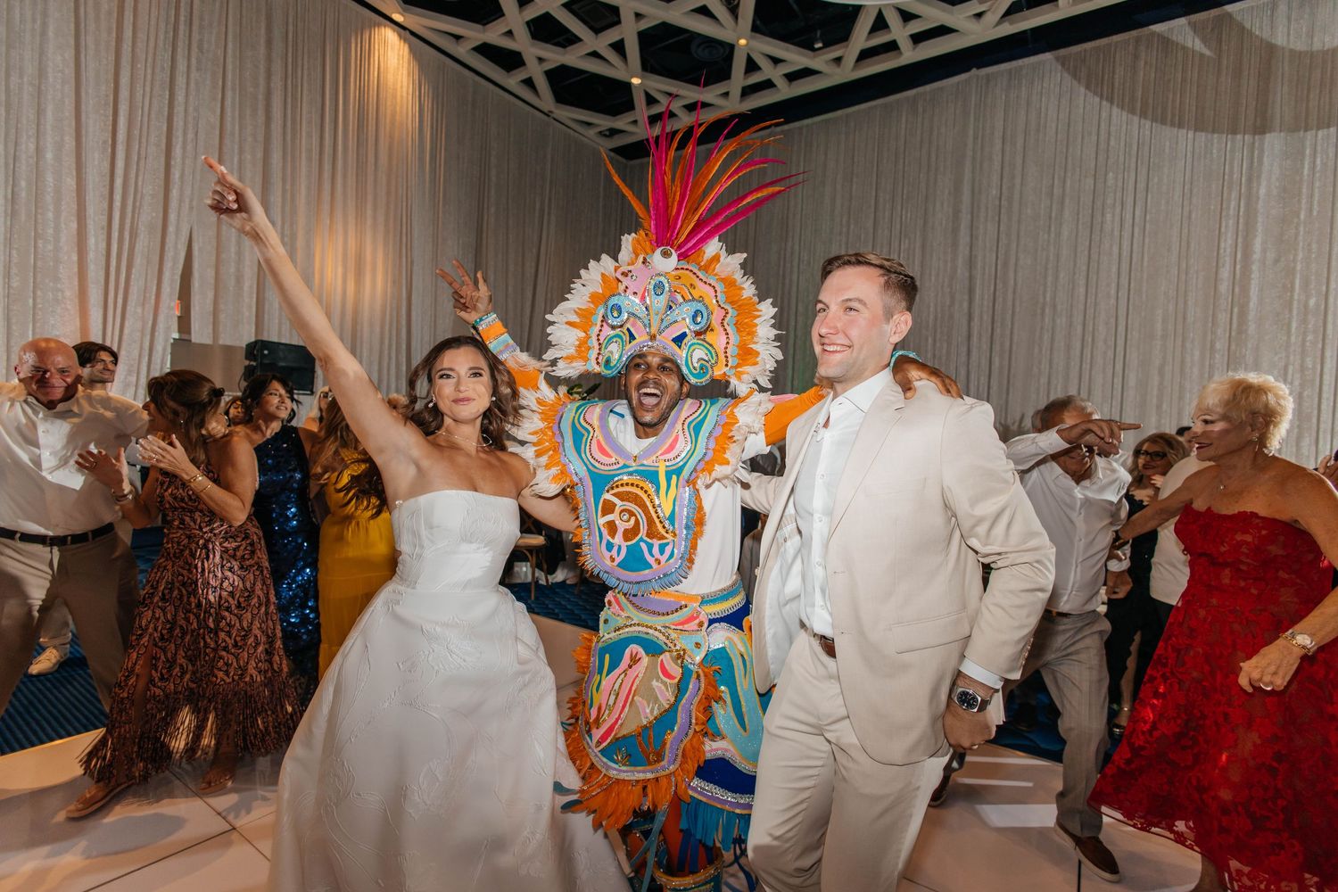 Bride and groom celebrate with colorful Junkanoo performer on wedding dance floor.