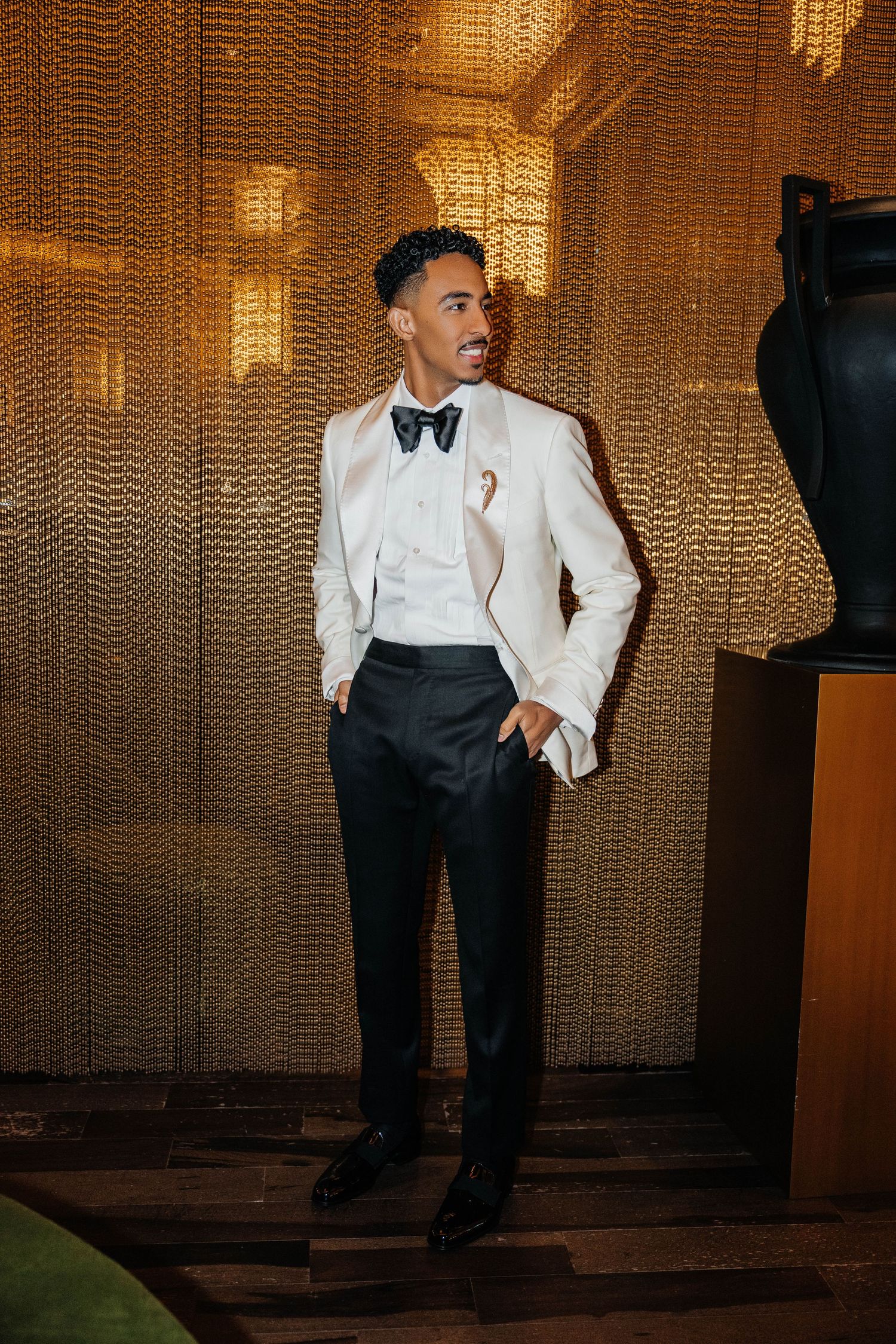 Stylish man in white tuxedo jacket, black bow tie, and trousers poses confidently against a golden textured wall.