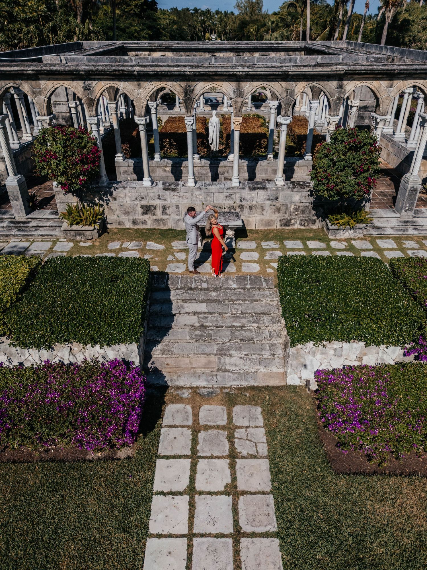 Stone pathway lined with purple flowers leads through a historic courtyard with arched columns and greenery.