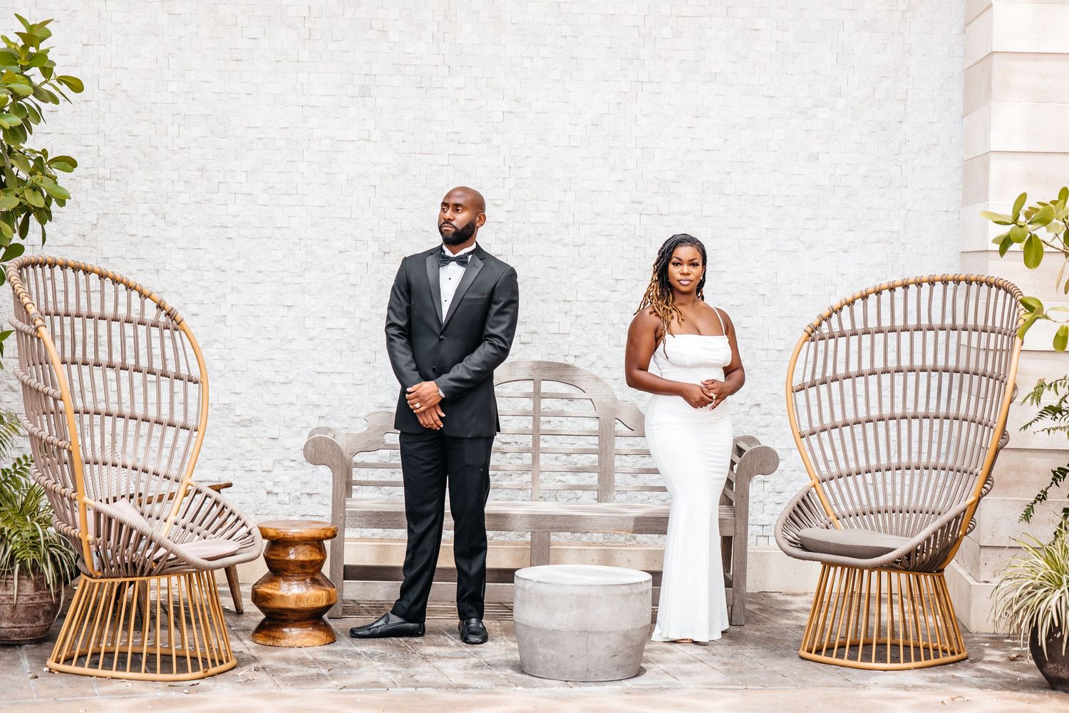 Elegant couple poses in formal attire against white wall with rattan chairs.