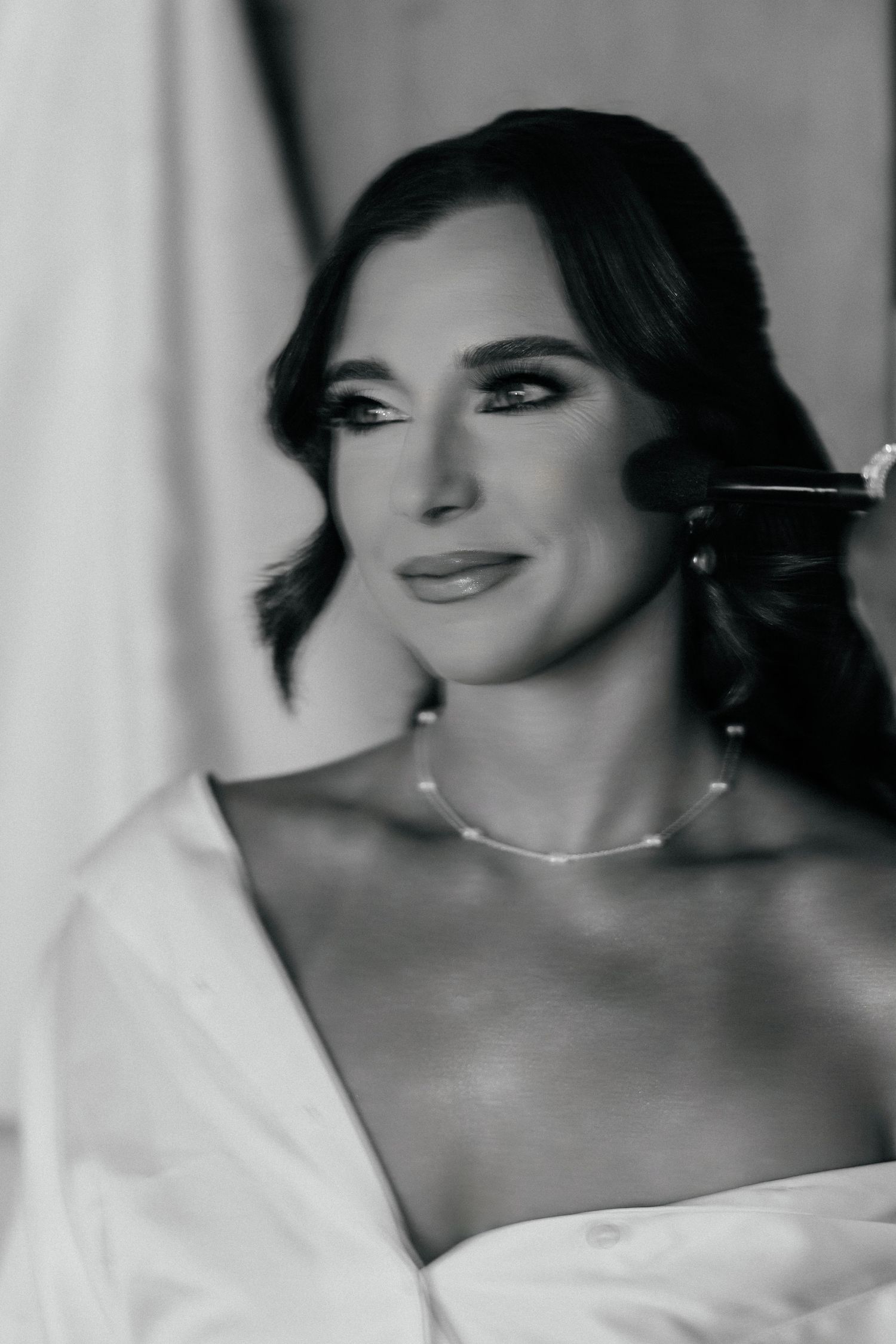 Black and white portrait of a bride in pearl necklace getting makeup applied, elegant wedding preparation photo.