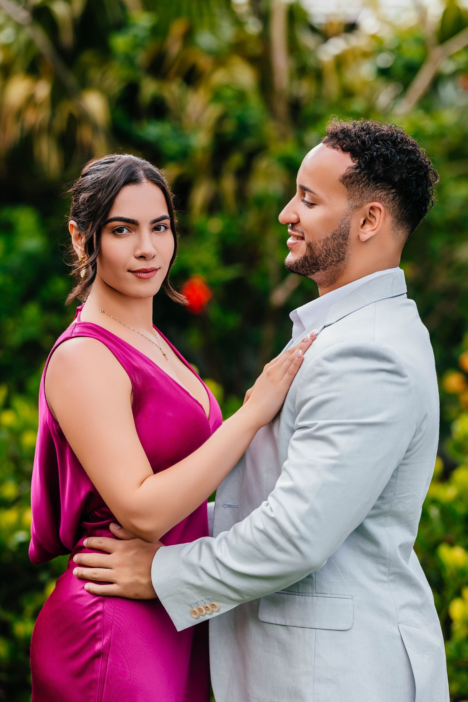 A formal couple embraces in pink and gray attire against tropical foliage.
