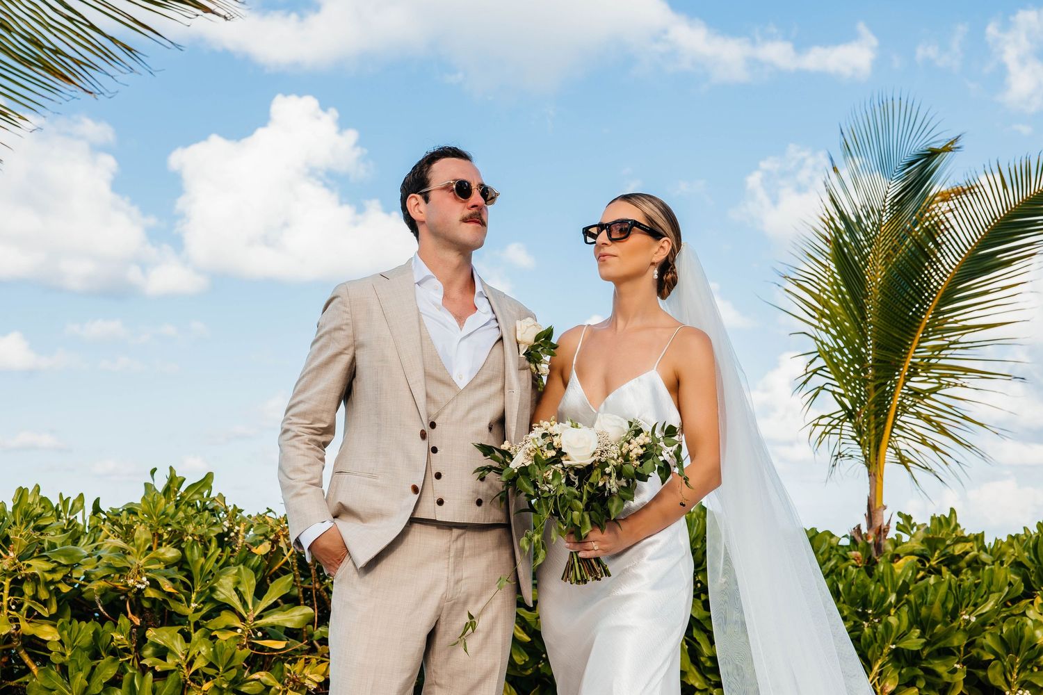 Beach wedding couple in sunglasses poses with tropical palm trees and greenery.