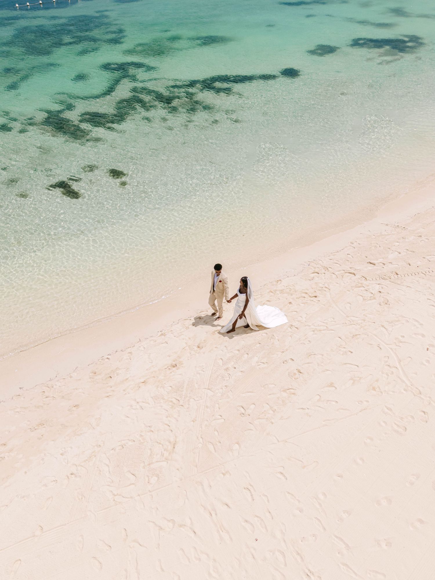 Aerial view of a couple in wedding attire holding hands on a pristine white sand beach with turquoise waters.