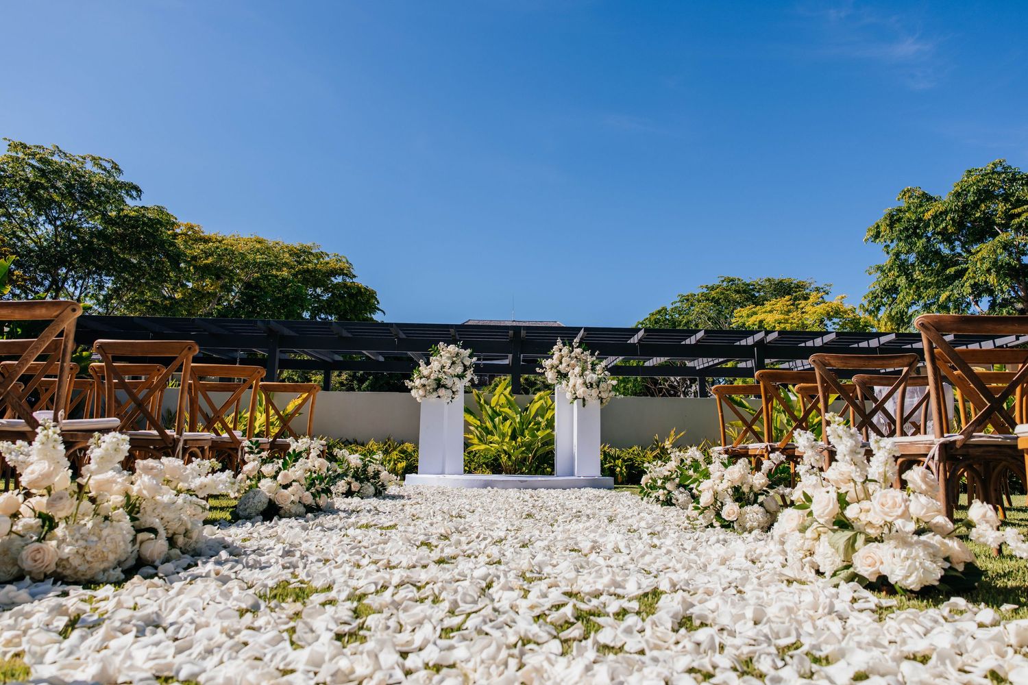 Elegant outdoor wedding ceremony aisle lined with white floral arrangements leading to a white altar under blue skies.