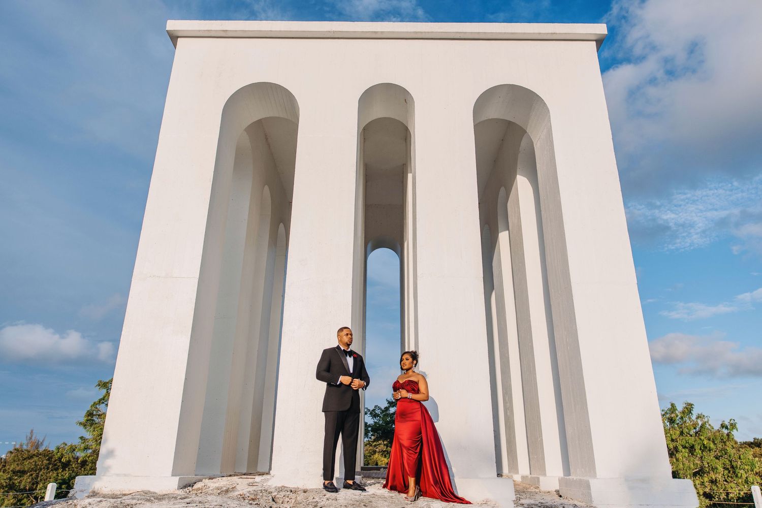 Elegant couple photographed under white architectural columns against blue sky.