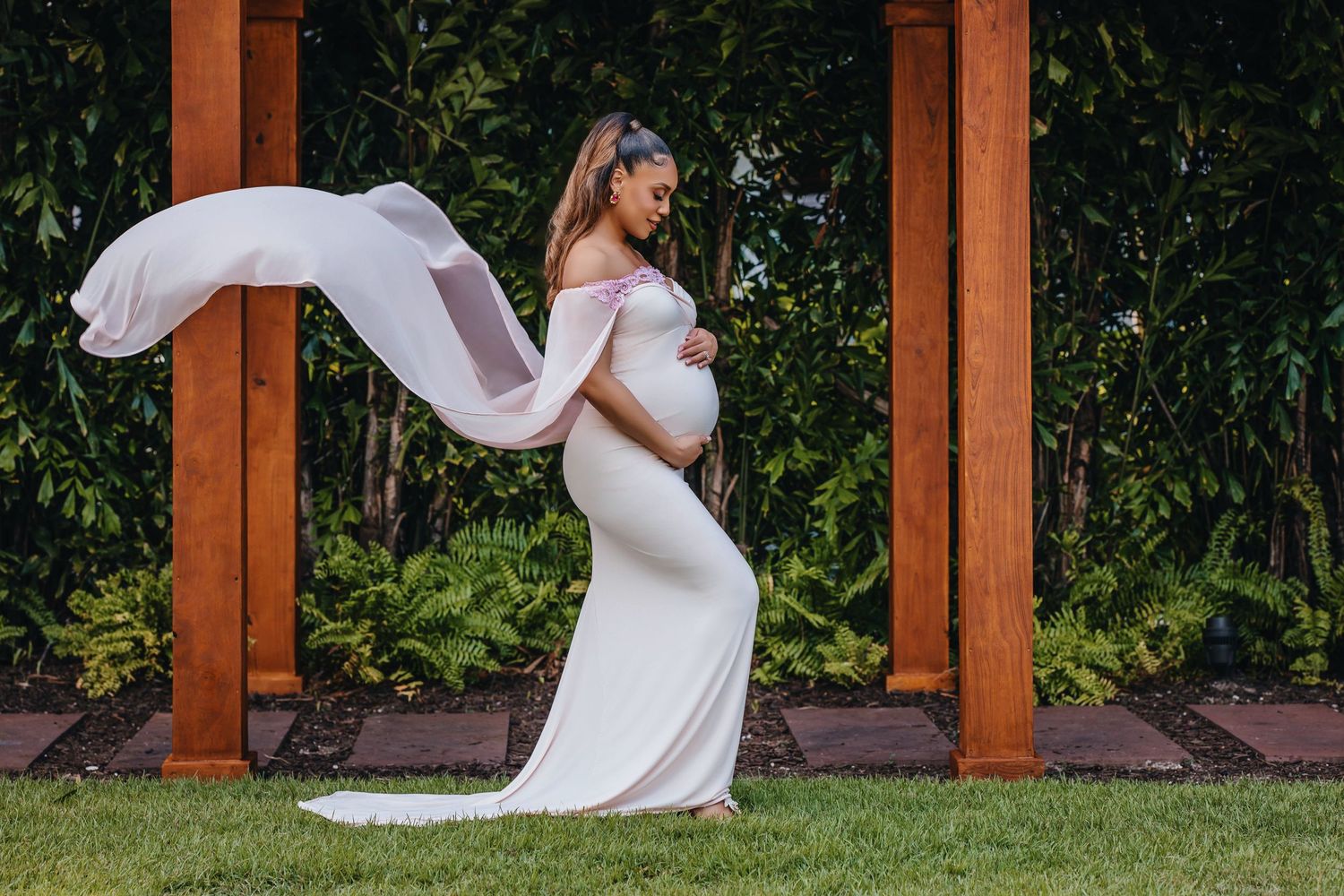 Maternity photo of a flowing white dress and cape against a wooden pergola backdrop.