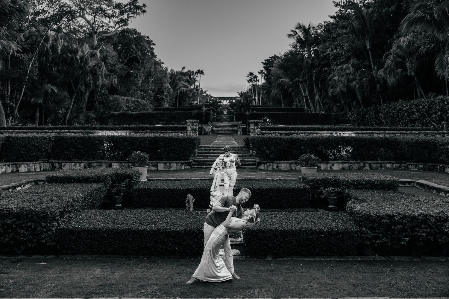 Black and white photo of couple in formal wear posing in garden setting.