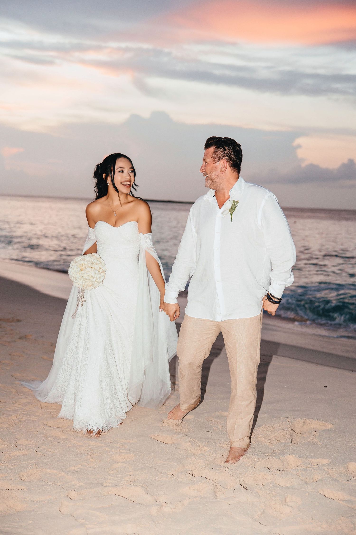 Bride and groom walk hand-in-hand on a sandy beach at sunset, bride in white gown holding bouquet.