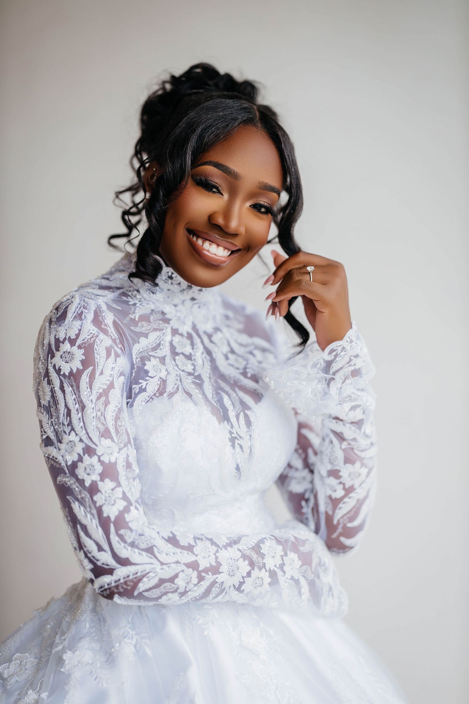 A radiant bride in a white lace long-sleeve gown smiles beautifully, showcasing elegant bridal makeup and styled curls.