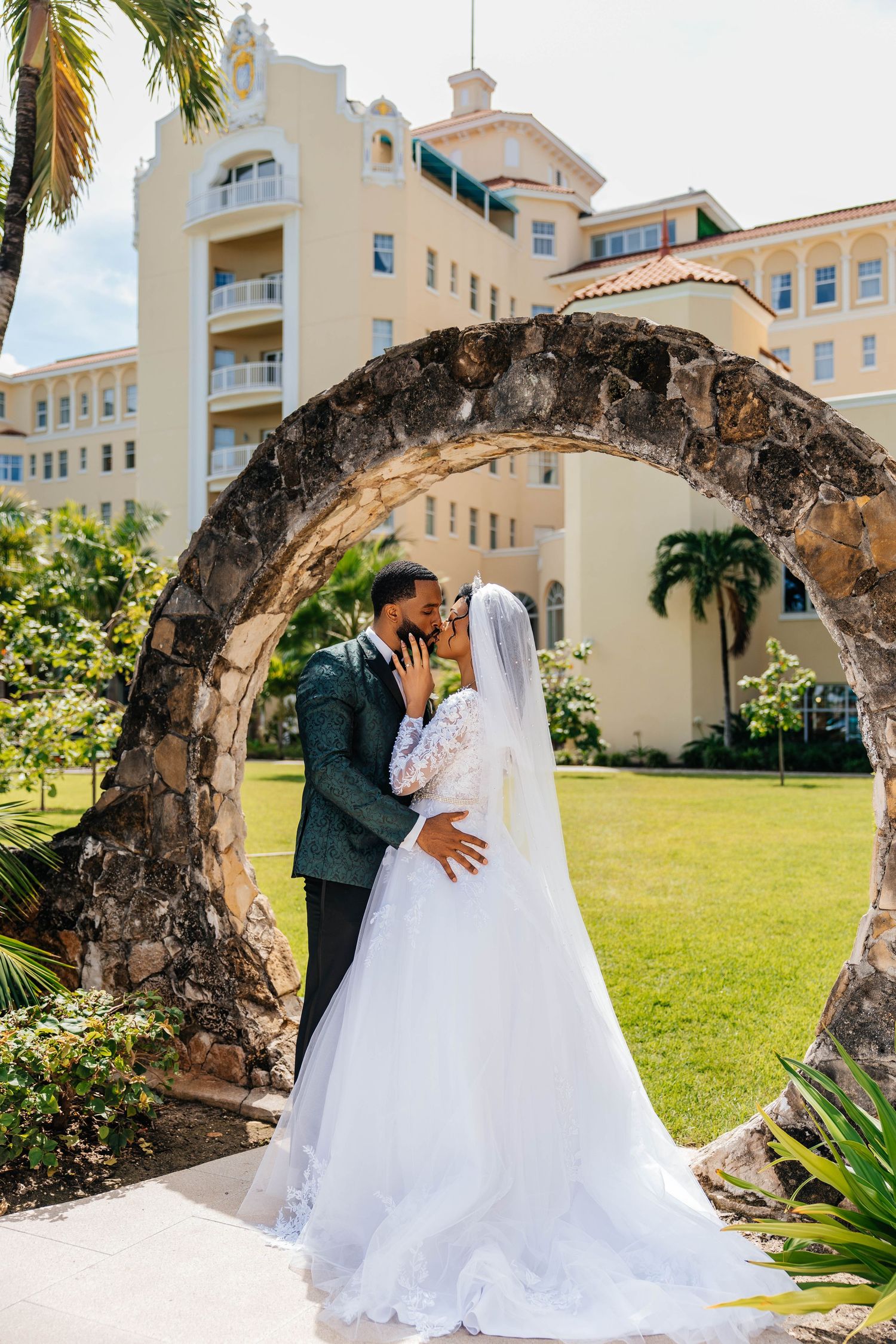 A couple shares a romantic moment under a stone archway at a tropical resort with palm trees and Spanish architecture.