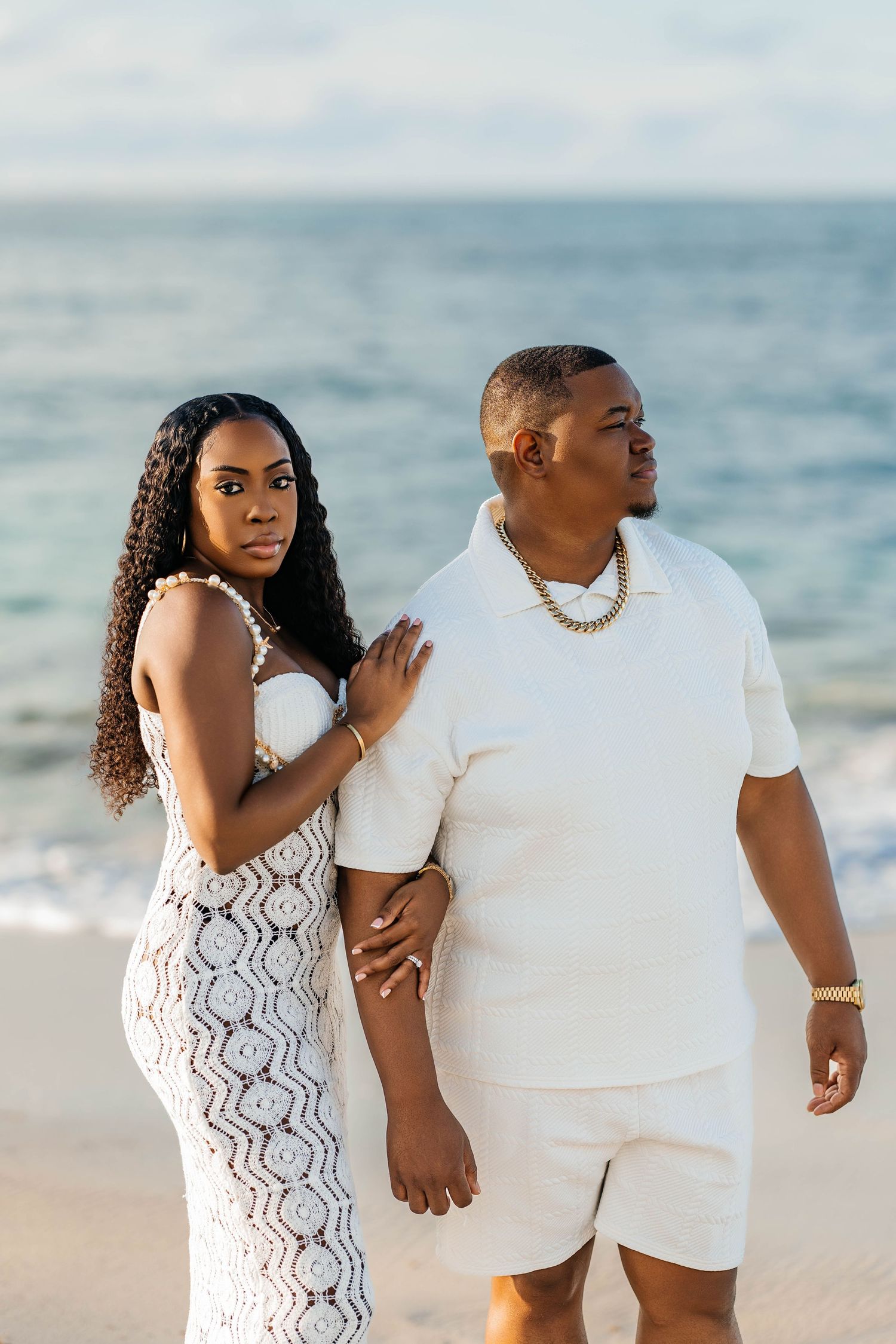 A couple in coordinating white outfits shares intimate moments during a romantic beach photoshoot at sunset.