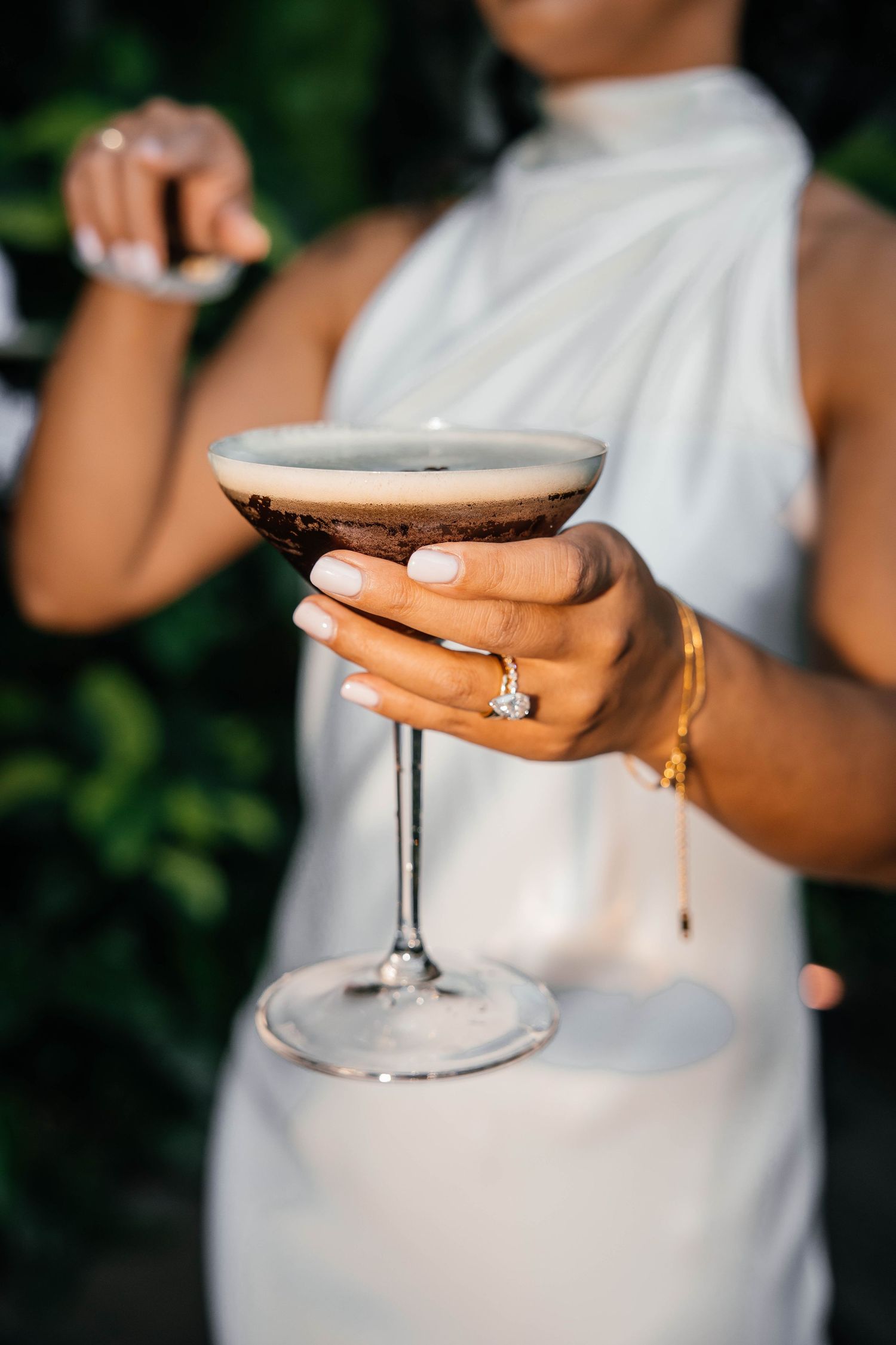 Bride holding an espresso martini cocktail in a coupe glass, wearing a diamond ring and gold bracelet in a white dress.