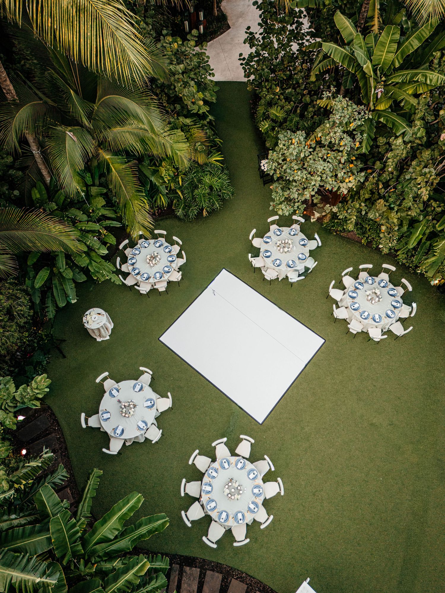 Aerial view of elegant outdoor wedding reception with round tables on green lawn surrounded by tropical foliage.
