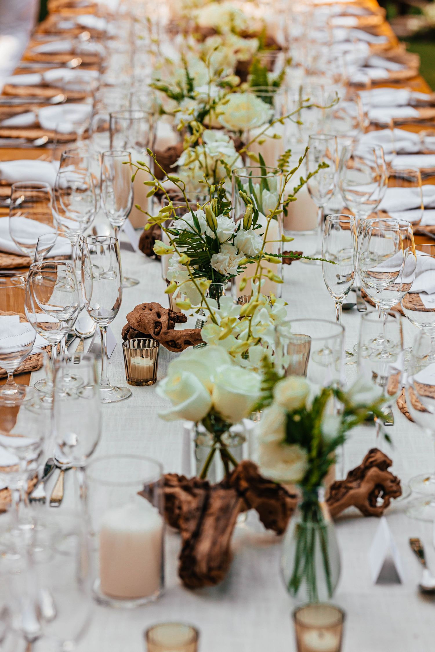 Luxurious wedding reception table set with white roses, driftwood centerpieces, crystal glassware, and candles on white linen.