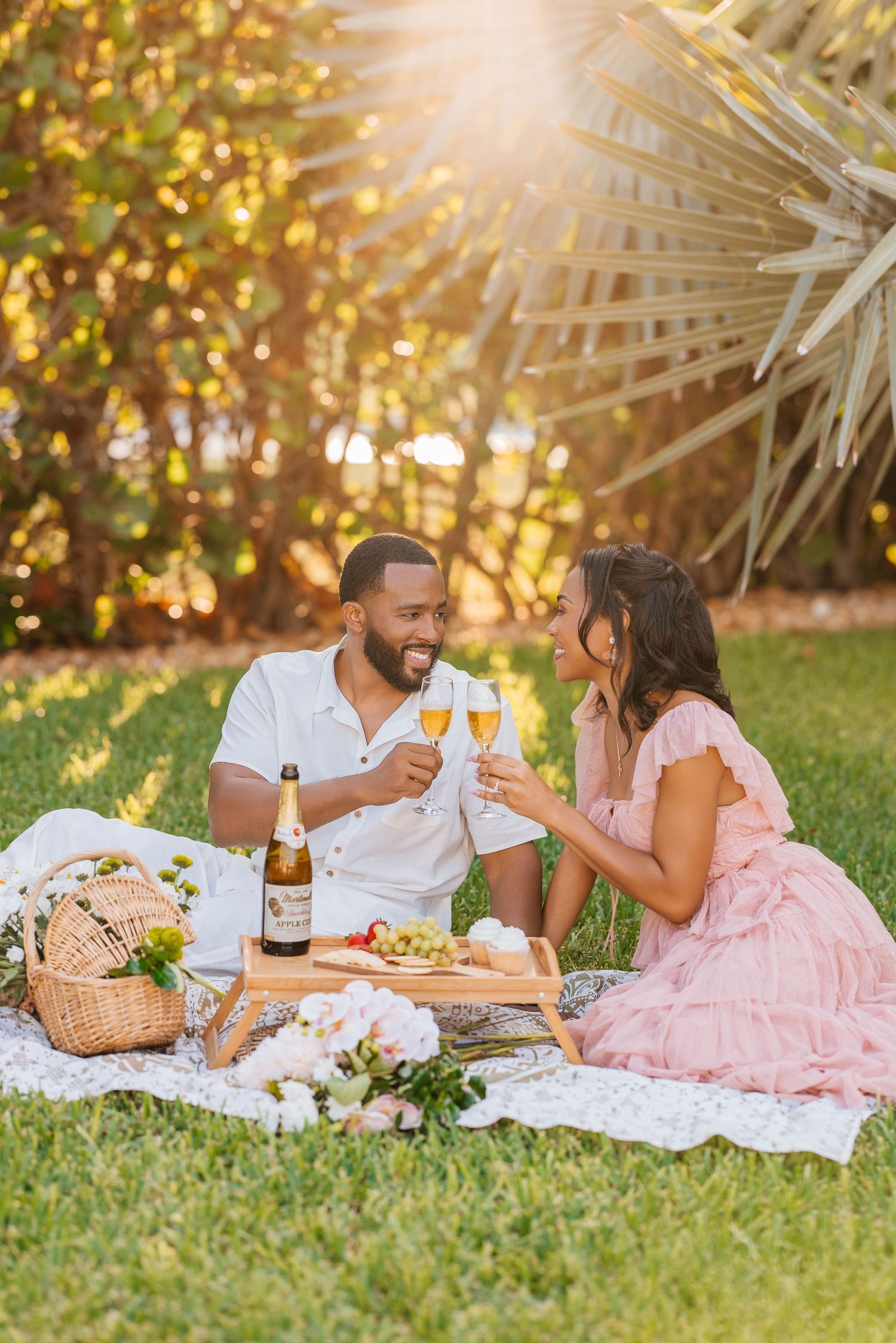 A romantic picnic on a blanket with wine and food in a sun-dappled garden with palm trees overhead.