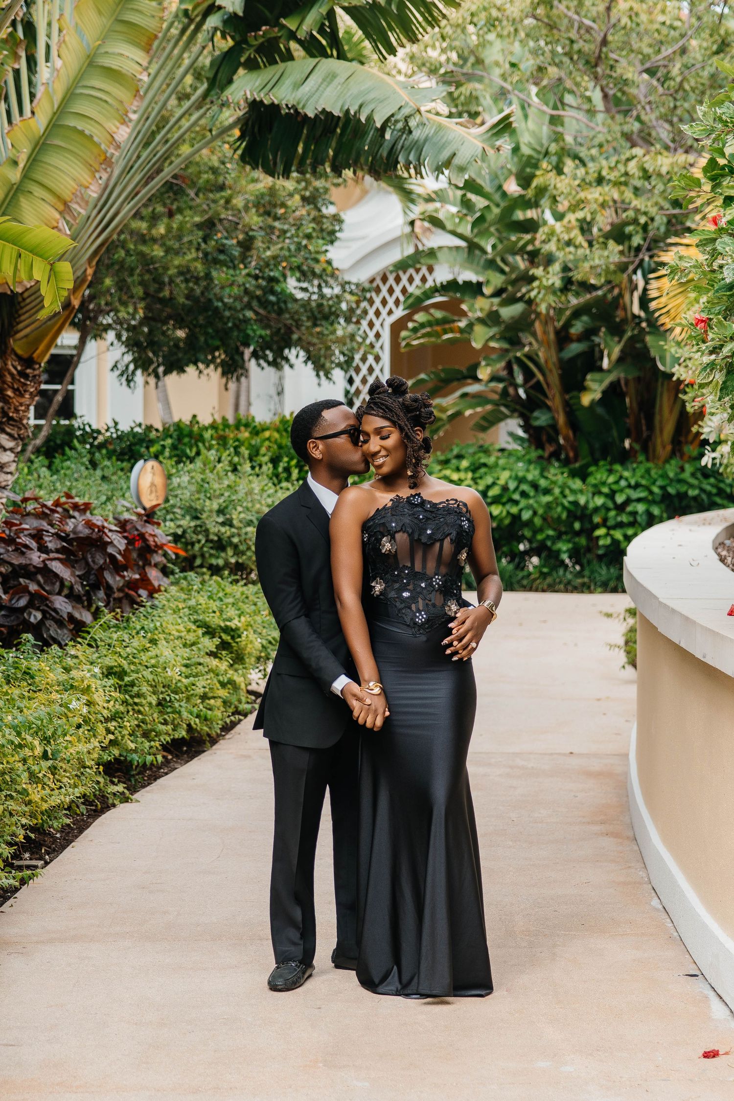 A couple in elegant black formal attire shares a romantic kiss in a lush tropical garden setting.