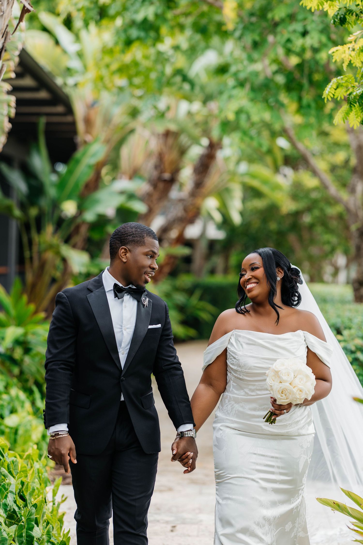 A joyful bride and groom walk hand in hand through a lush tropical garden, smiling at each other on their wedding day.