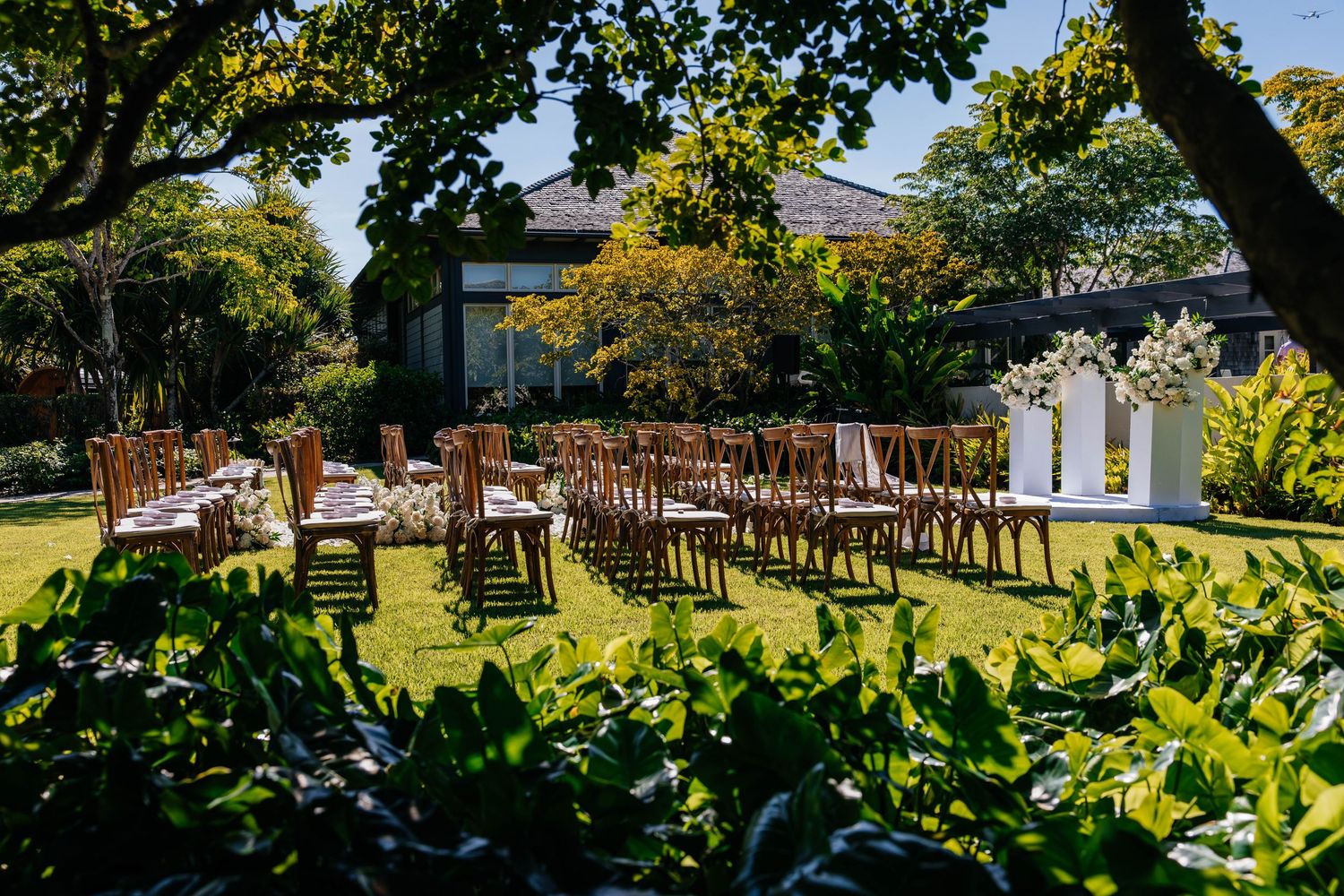 Outdoor wedding ceremony setup with wooden chairs and tables on lush green lawn surrounded by tropical trees and garden.